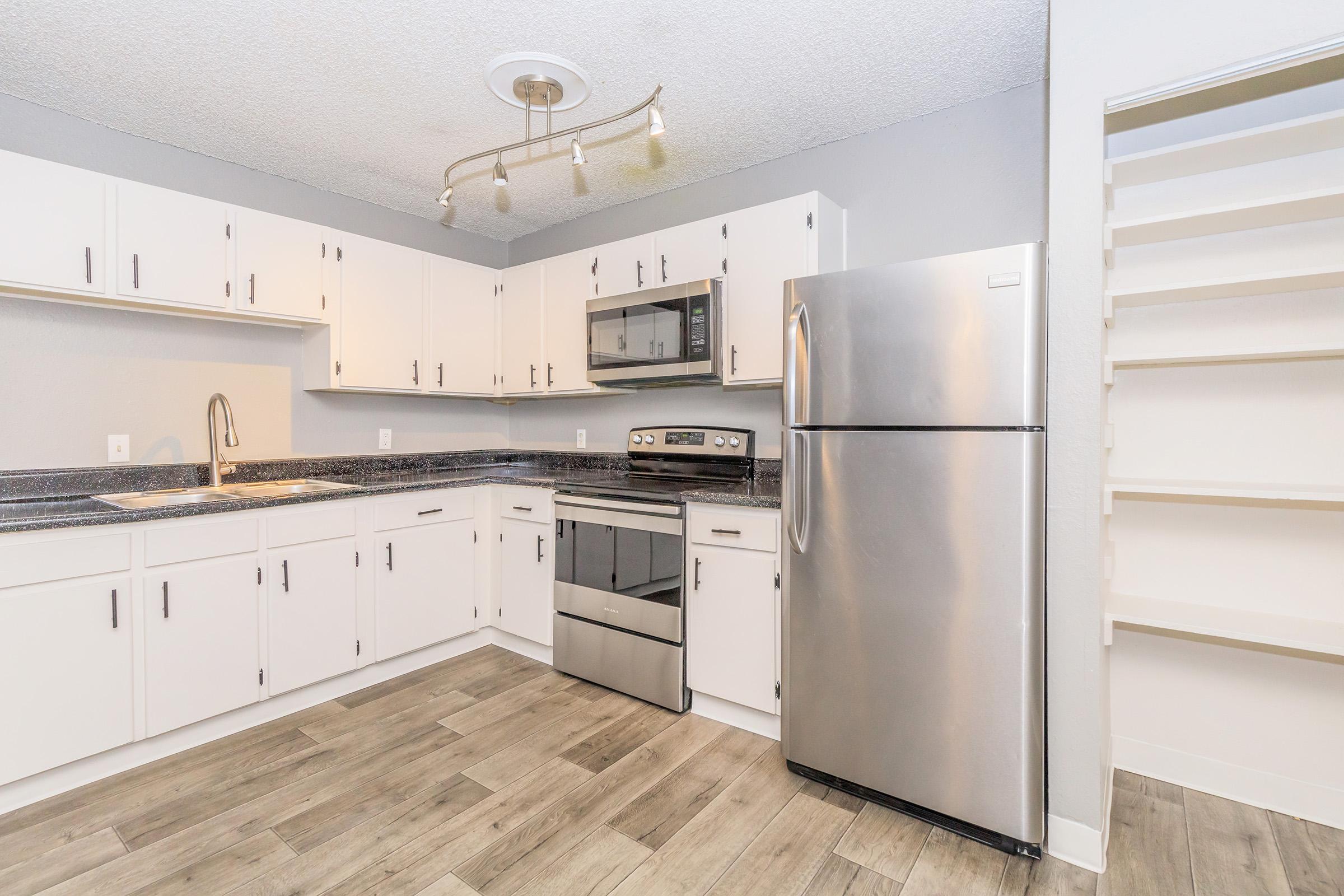 Modern kitchen featuring white cabinetry, stainless steel appliances including a refrigerator and oven, and dark countertops. A track light illuminates the space, complemented by light-colored wood flooring. Shelving is visible on the right side, providing additional storage options.