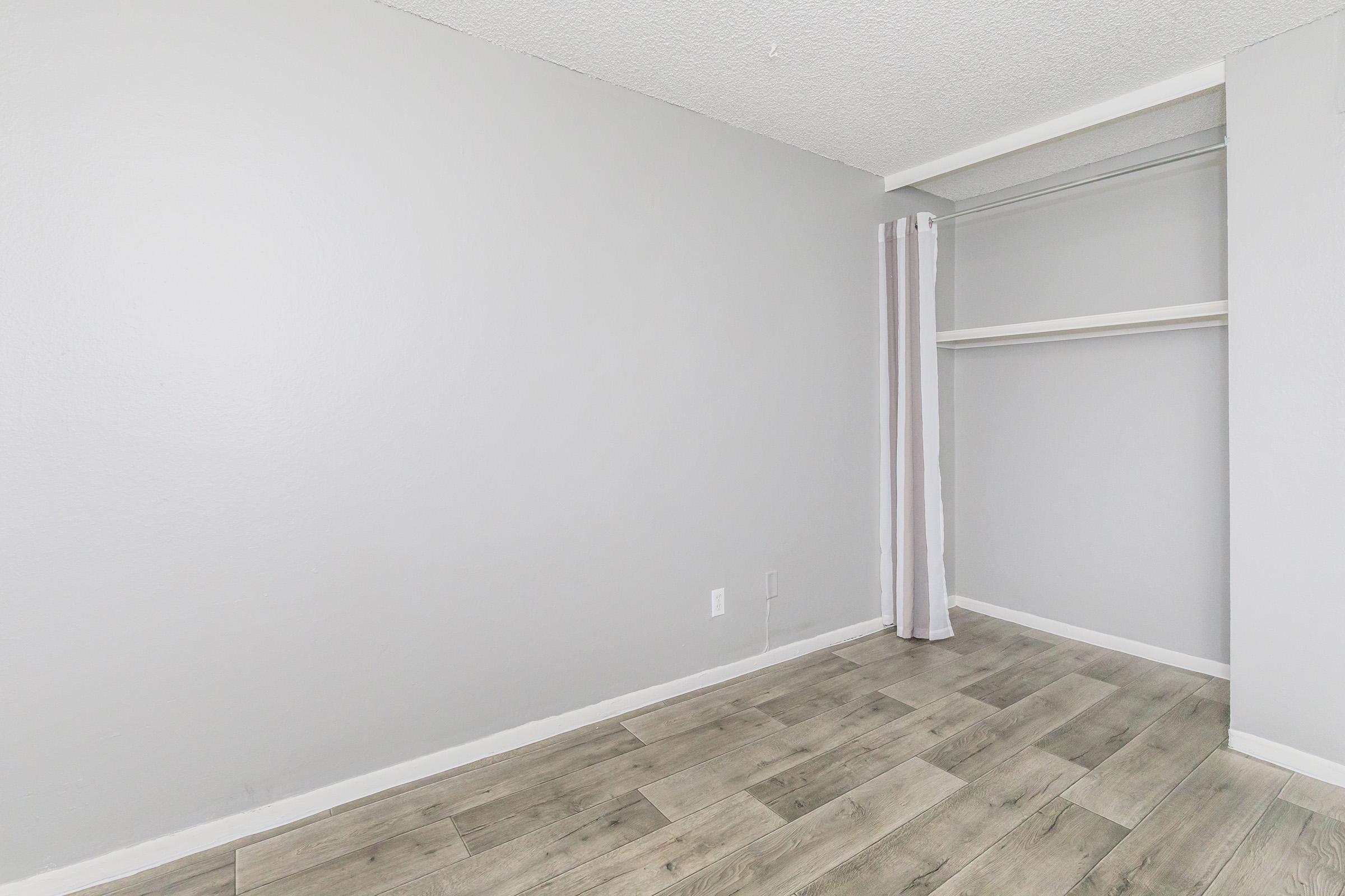 A minimalistic room featuring light gray walls and a wood-style floor. On one side, there is a closet with an open shelf and a white curtain partially covering the entrance. The space is empty, giving it a clean and uncluttered look.