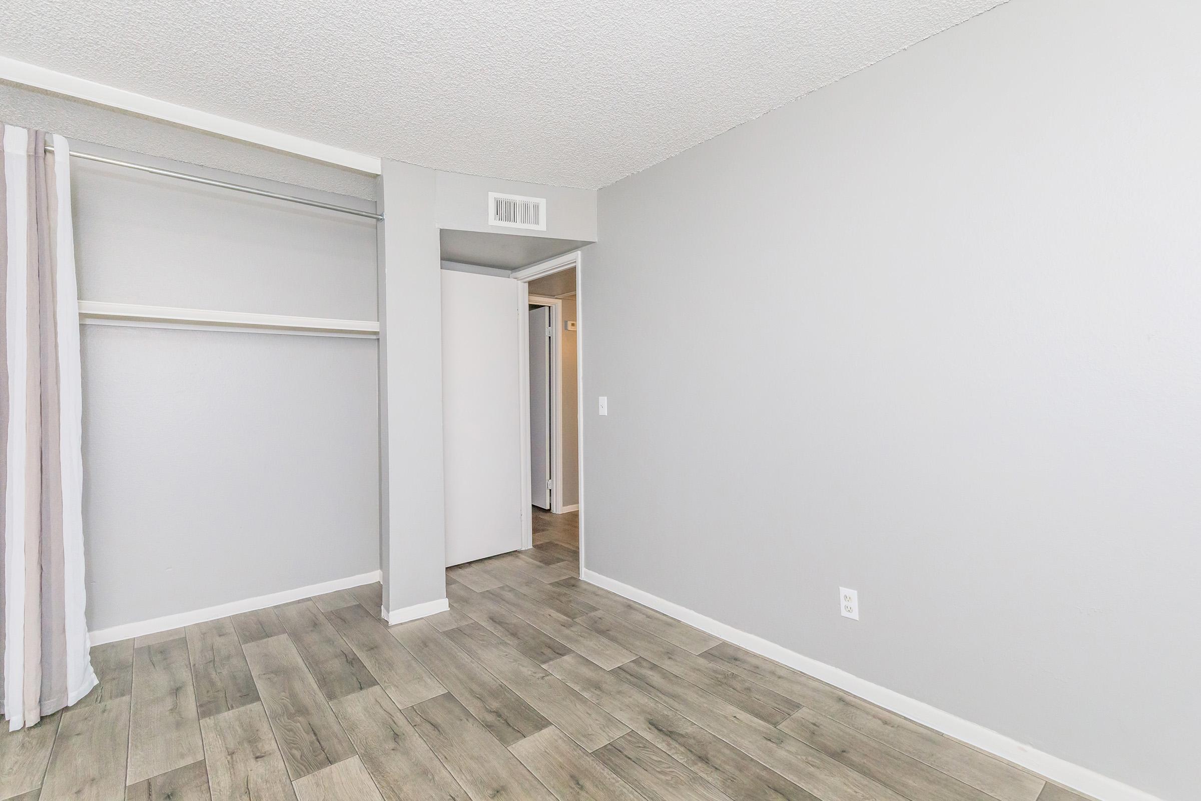 An empty room featuring light gray walls, a closet with a hanging rod, and a doorway leading to another space. The floor is composed of light-colored wood planks, and there is a curtain hanging on one side of the room. The overall atmosphere is bright and spacious.