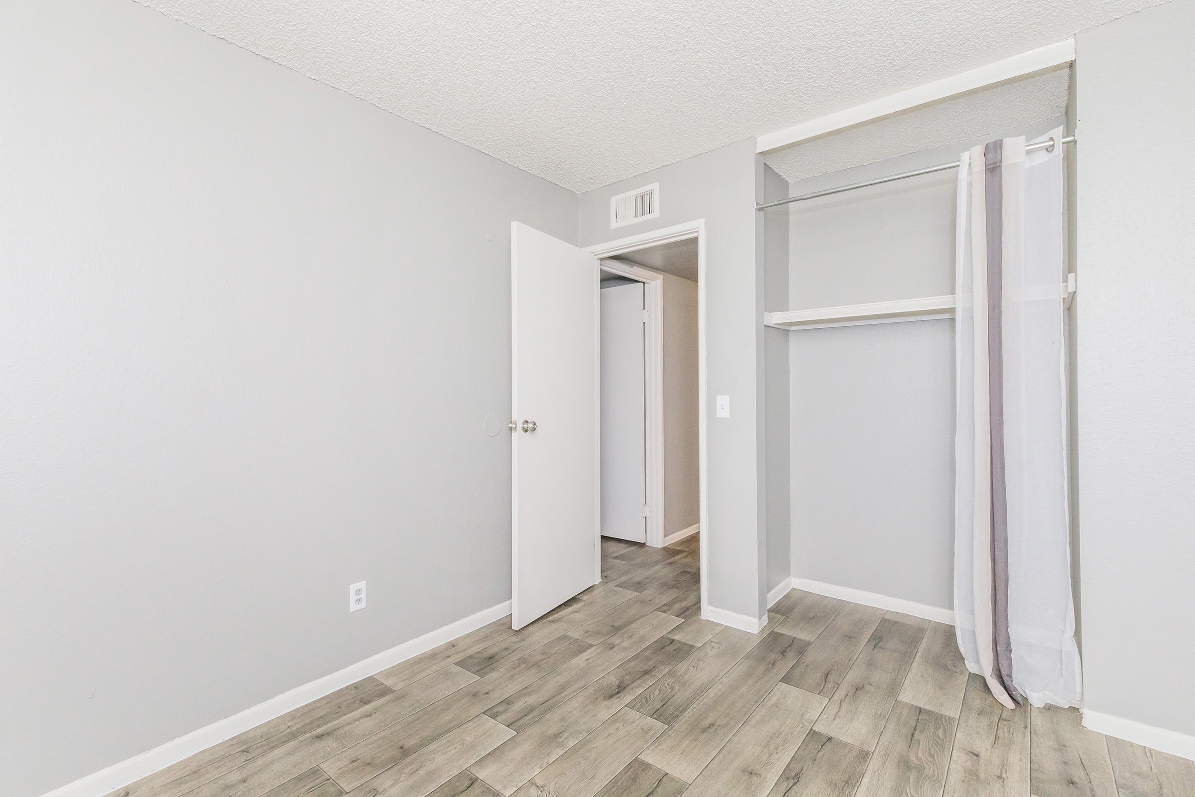 A small, empty room with light gray walls and a wood-look floor. There is a white door slightly ajar on the left, and a built-in closet with a hanging rod on the right. A sheer curtain hangs in front of the closet. The room is well-lit and has a clean, minimalist appearance.