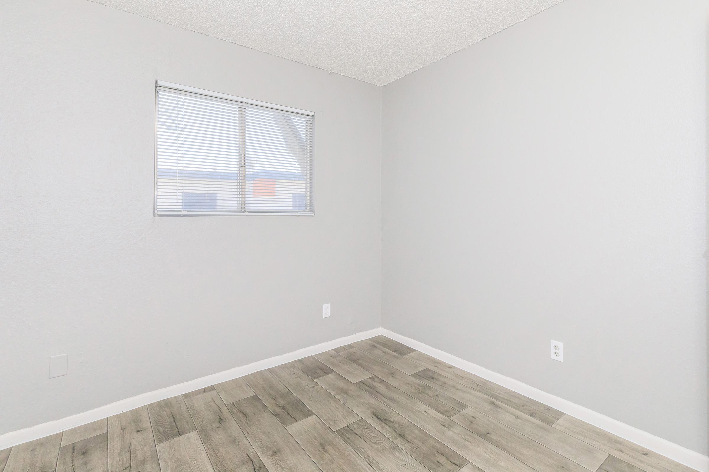 A vacant room with light gray walls and light wood-like flooring. There is a window with blinds on the left side, letting in natural light. The room is empty, creating a clean and minimalistic appearance, suitable for various purposes such as an office or bedroom.