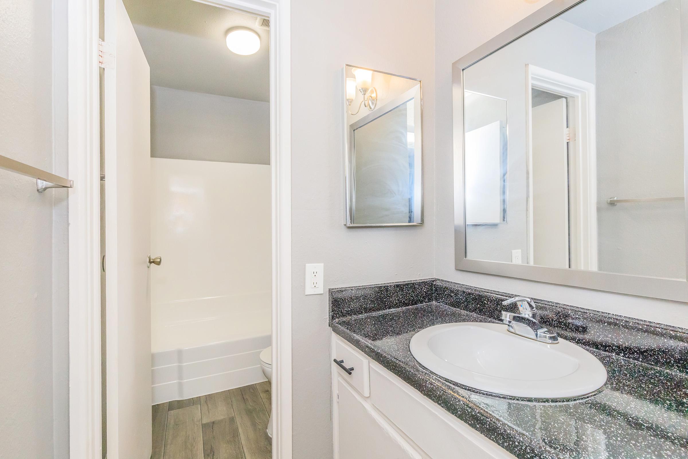 Modern bathroom featuring a sleek countertop with a round sink, a large mirror above, and a light fixture. A door leads to a shower area with a white tub and light-colored walls, enhancing the spacious look with gray flooring. The overall design is clean and contemporary.