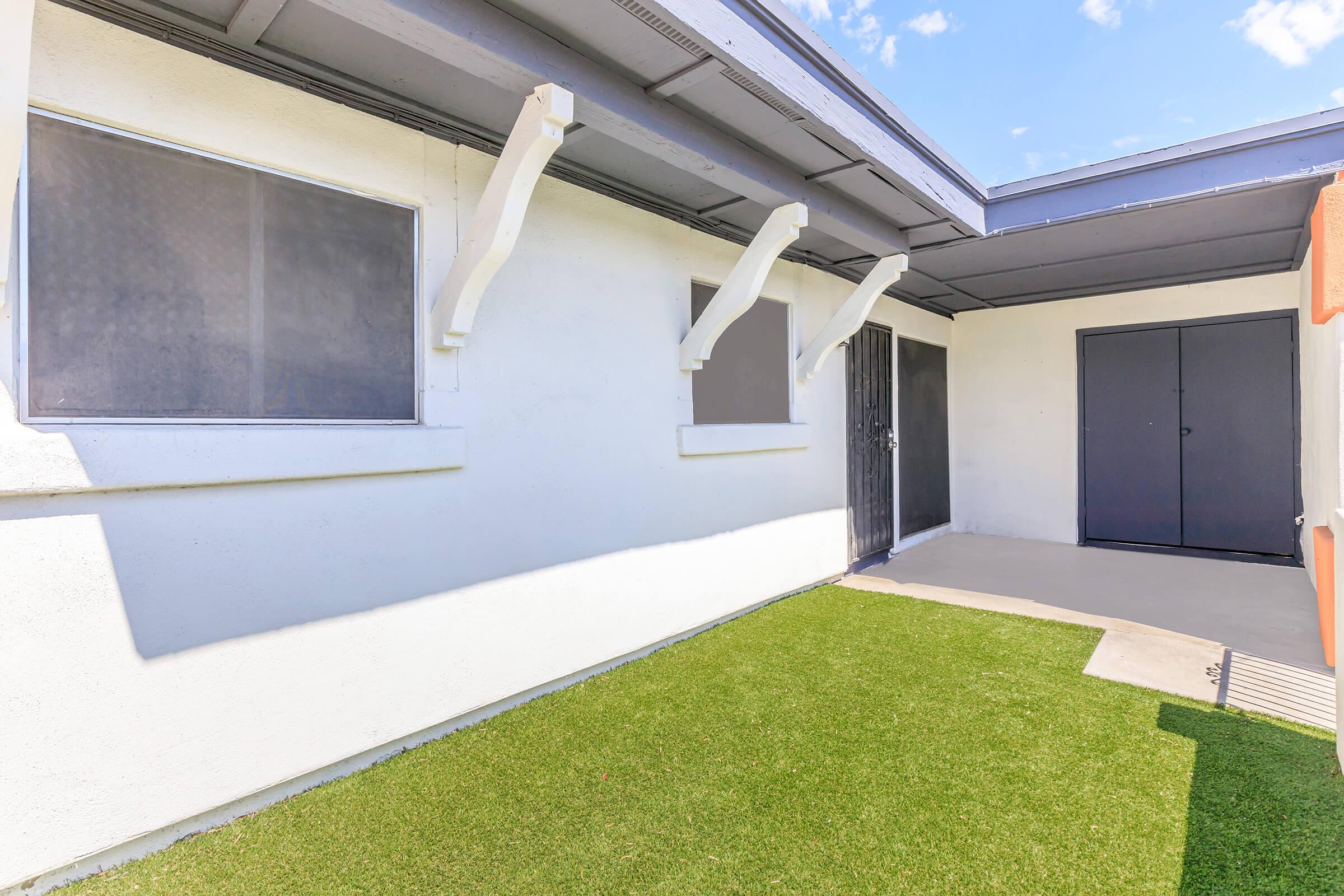 A view of a small outdoor patio area featuring artificial grass, with two windows and a black door visible. The patio is bordered by white walls and has a covered section overhead. The sky is clear with a few clouds.
