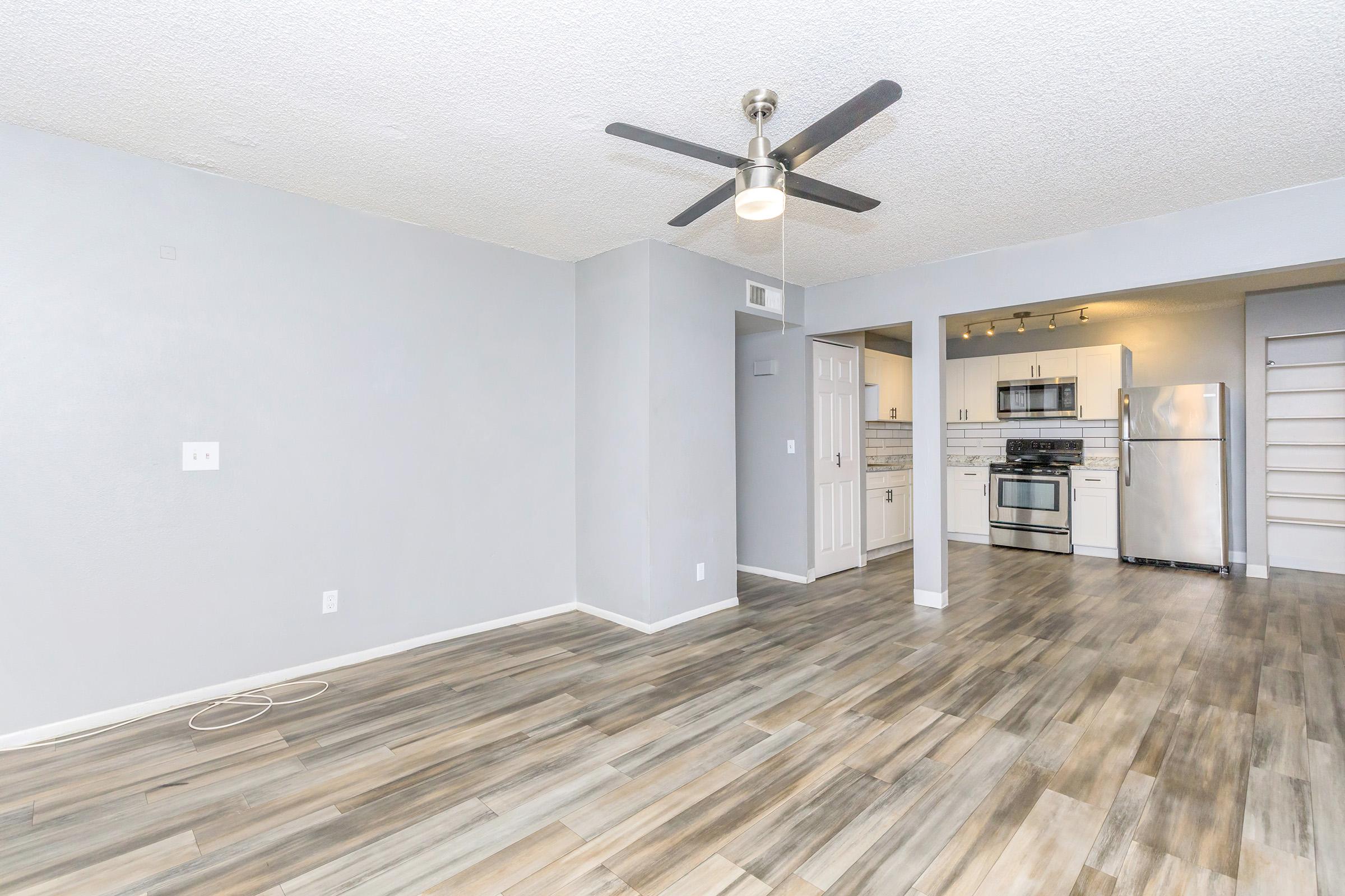 Open living space featuring gray walls and a ceiling fan. The room has wood-like flooring and connects to a modern kitchen with white cabinets, stainless steel appliances, and a countertop. Bright lighting enhances the inviting atmosphere, making it suitable for relaxation or social gatherings.