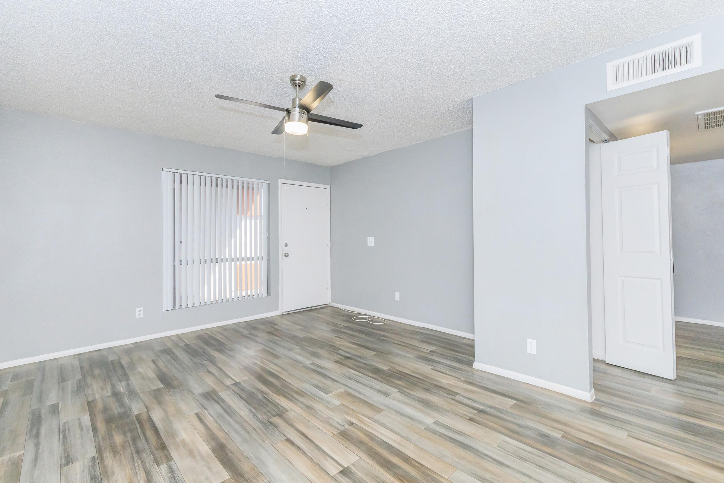 Spacious living area featuring light gray walls, a ceiling fan, and large windows with vertical blinds. The floor is covered in modern, light-colored laminate planks. A white door leads outside, and there is a doorway to another room on the right. The overall ambiance is bright and inviting.