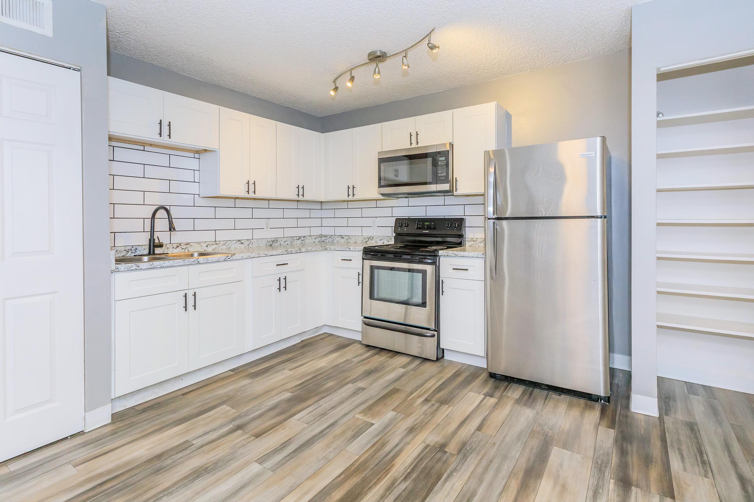 Modern kitchen featuring white cabinetry, stainless steel appliances, and a gray tile backsplash. The space includes a stovetop, microwave, and refrigerator, with a stylish countertop and wood-like floors. Natural light highlights the open layout and organized shelving.