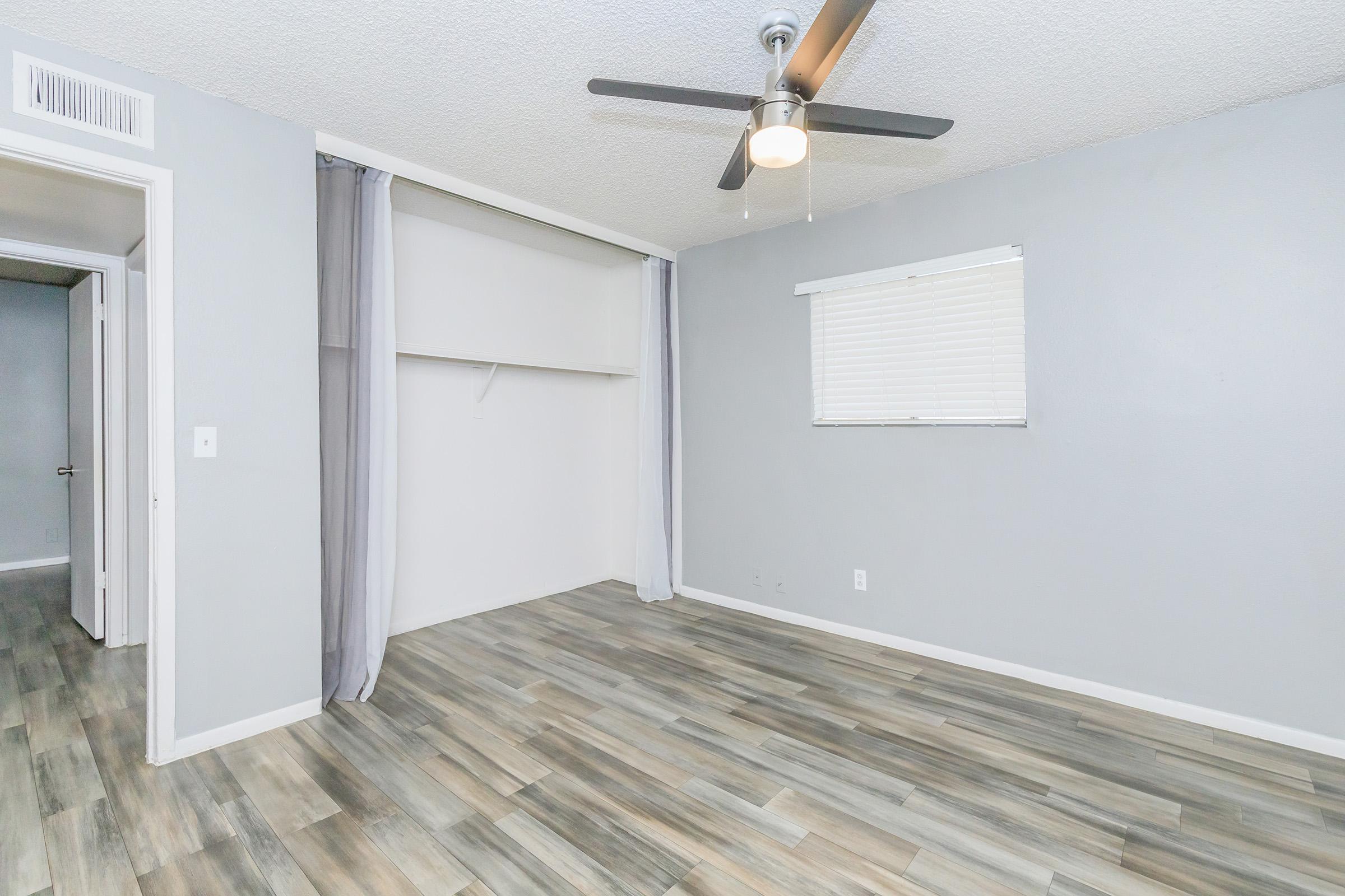 A spacious room featuring light gray walls and a ceiling fan. The floor is covered with modern, wooden-style tiles. There is a single window with blinds, and a small closet area is partially visible. Light curtains hang next to the closet, enhancing the room's natural brightness.