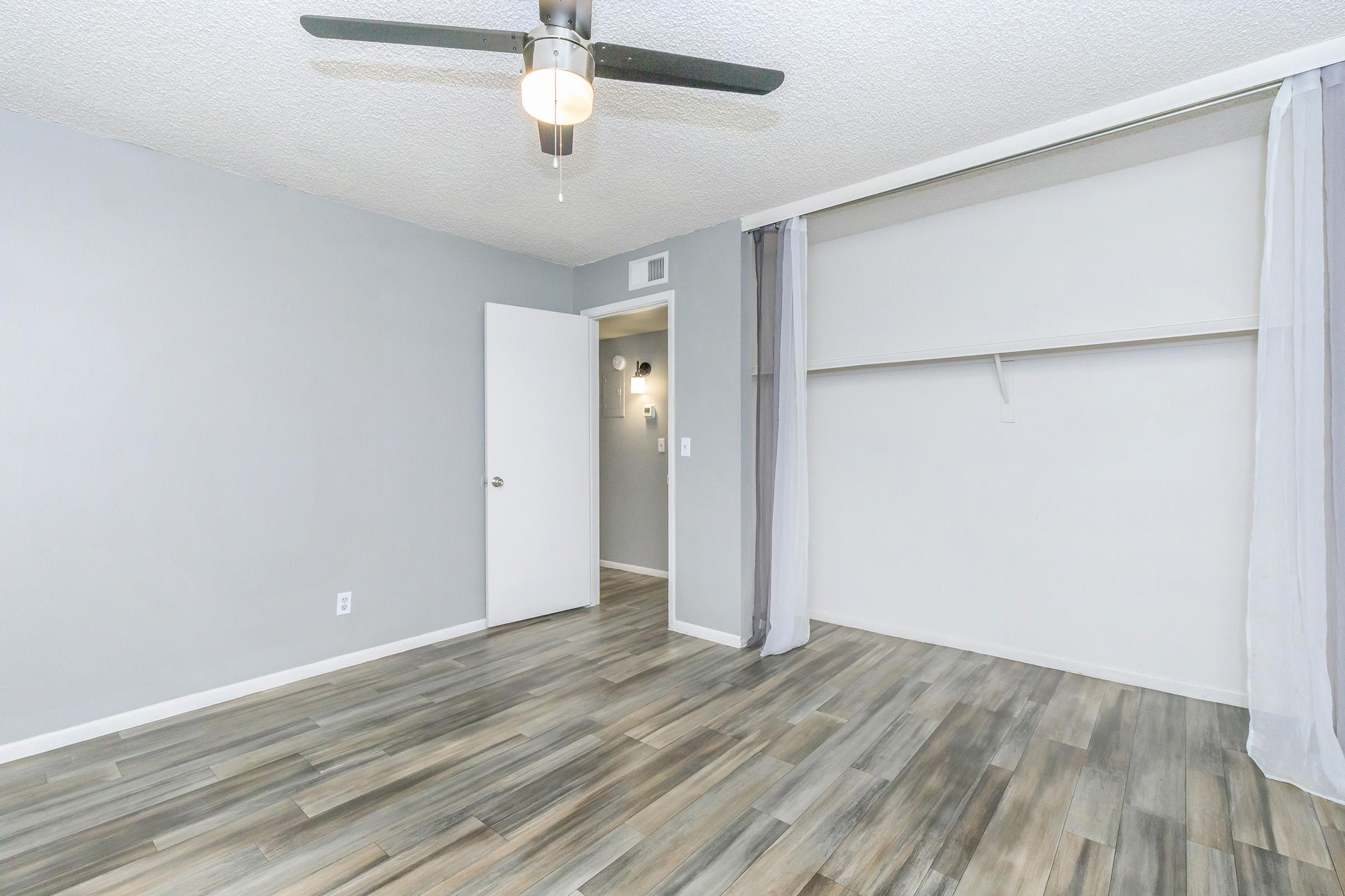 A modern, empty room featuring light gray walls, a light-colored wooden floor, a ceiling fan with a light fixture, and a closet with sliding doors. There is a white door leading to a small hallway in the background, along with sheer curtains on one side.