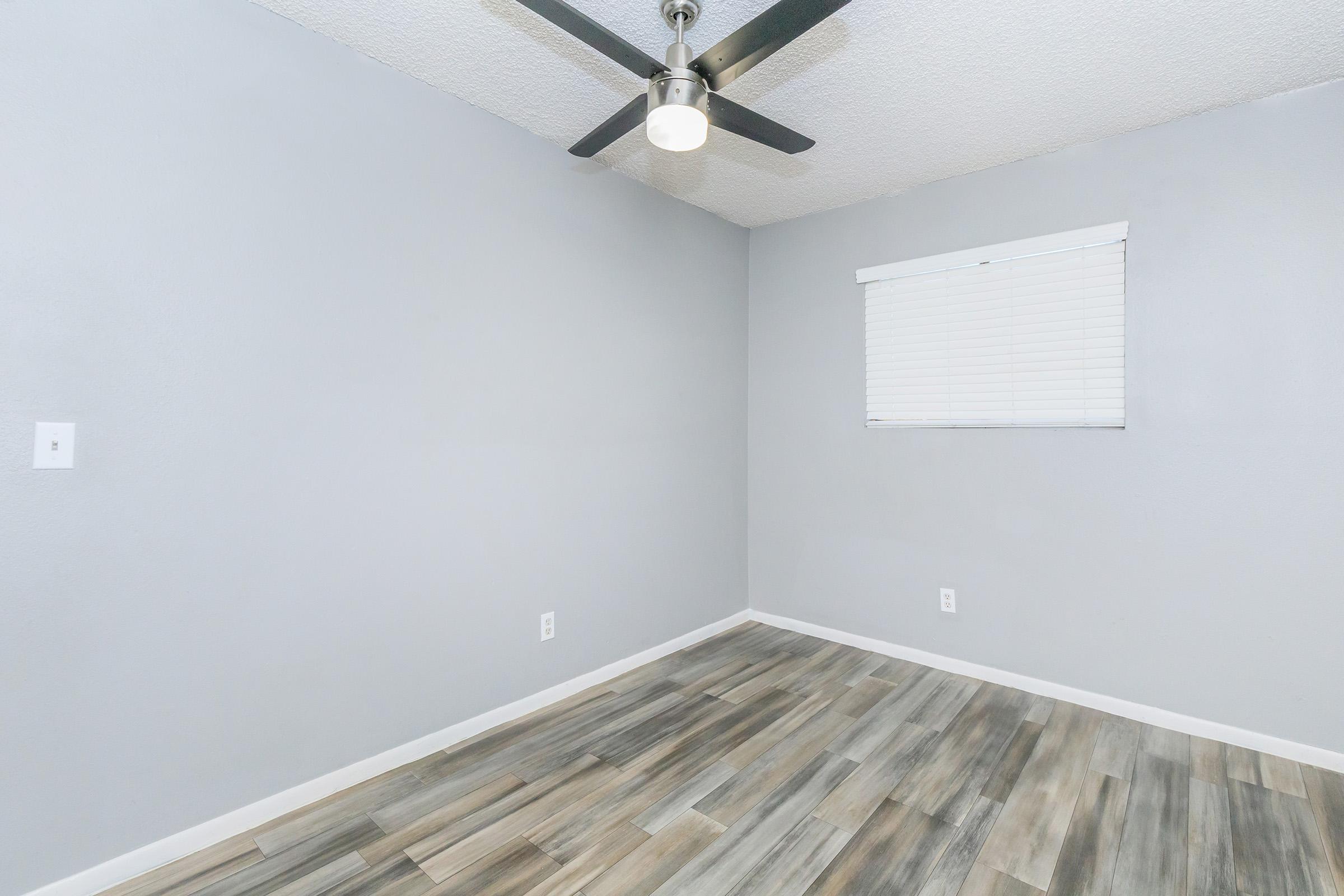 A minimalist room featuring light gray walls, a ceiling fan with a light fixture, a small window with blinds, and textured flooring in a mix of gray and beige tones. The space is empty, creating an open and airy feel.