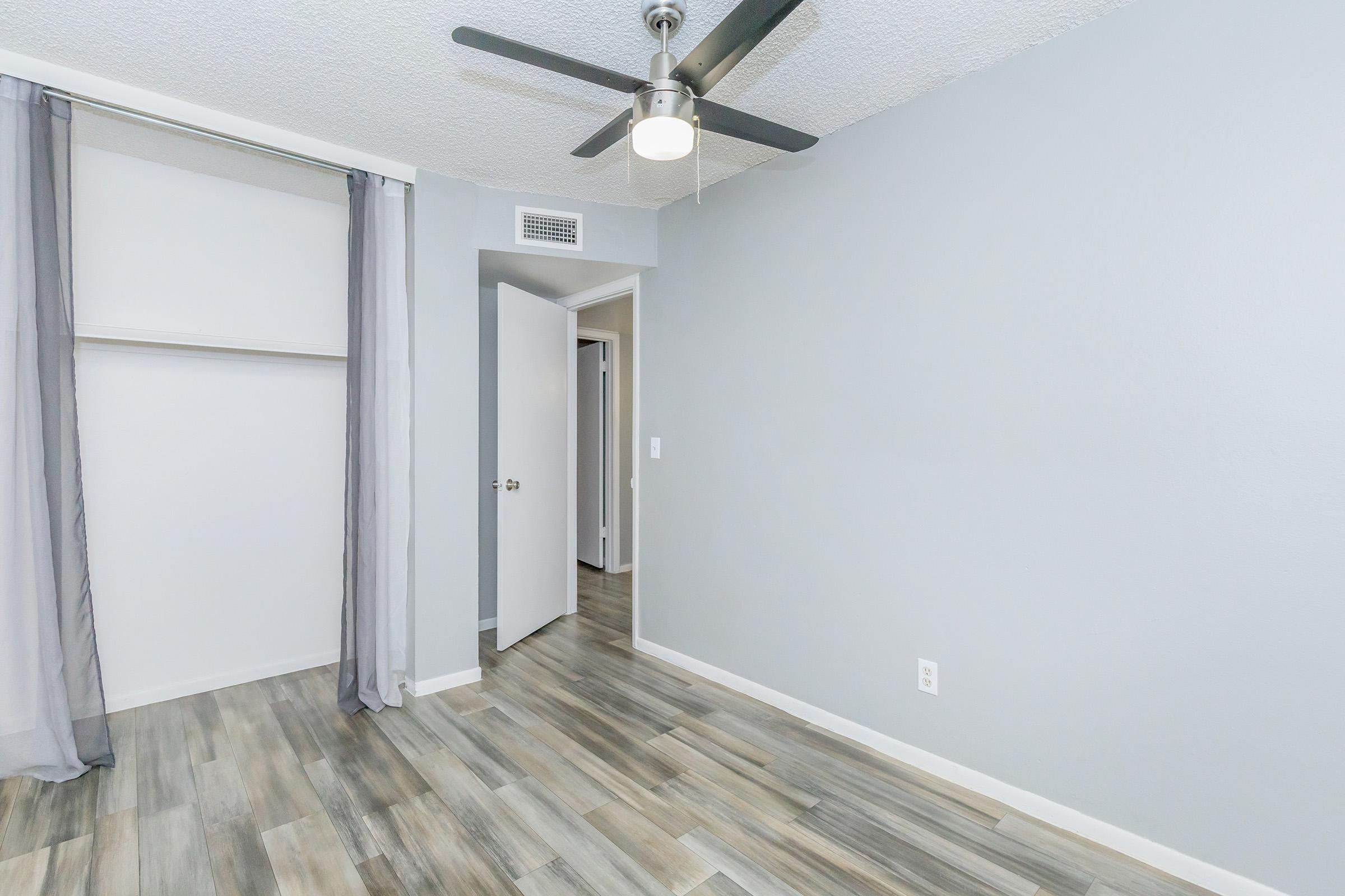 An empty room with light gray walls and a ceiling fan. The floor is covered with light wood-like tiles. There is a partially open closet on the left side with curtains, and a doorway at the back leading to another room. The space is bright and minimalistic, creating a clean aesthetic.