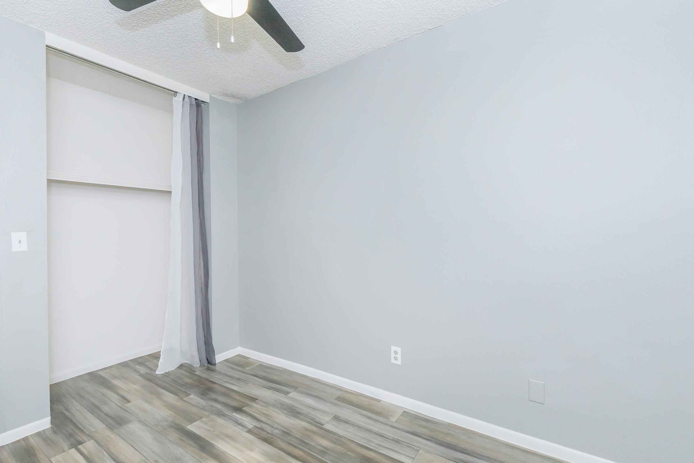 A spacious, empty room with gray walls and a ceiling fan. There is a light wooden floor and a sliding closet door partially open, revealing a light-colored interior. A sheer gray curtain drapes over the closet, adding a soft touch to the minimalist aesthetic.