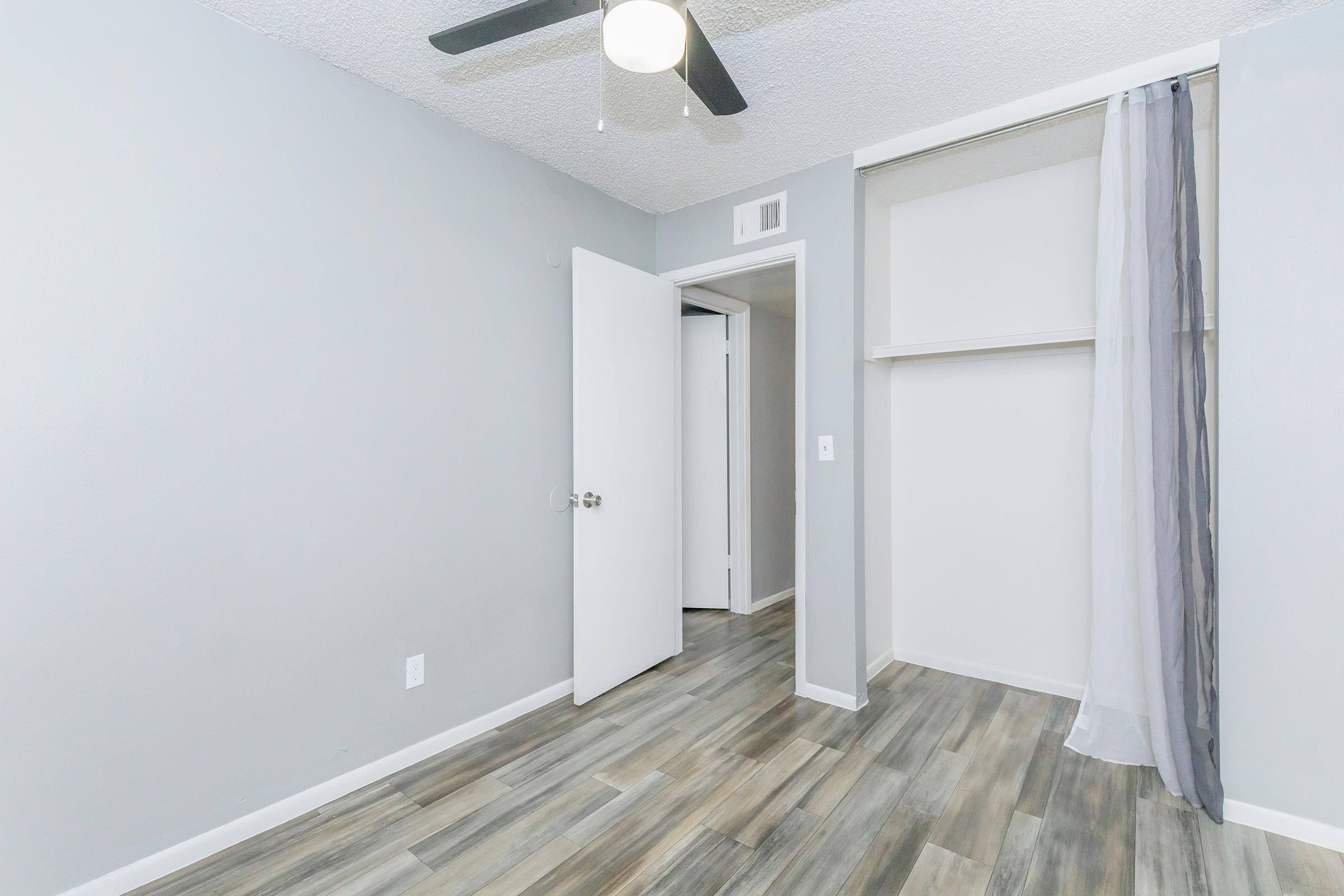 A well-lit interior room with light gray walls and wooden flooring. There is a white door partially open leading to another space, and a closet space with a curtain instead of a door. A ceiling fan is visible, providing a modern touch to the room.