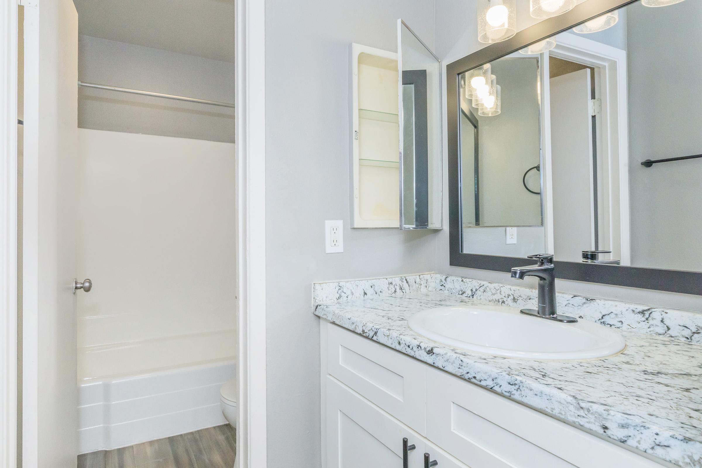 A modern bathroom featuring a white vanity with a granite countertop, a round mirror, stylish light fixtures, and a small shelving unit. A clean white bathtub is visible in the background, with light gray walls and wooden floorboards, creating a fresh and inviting atmosphere.