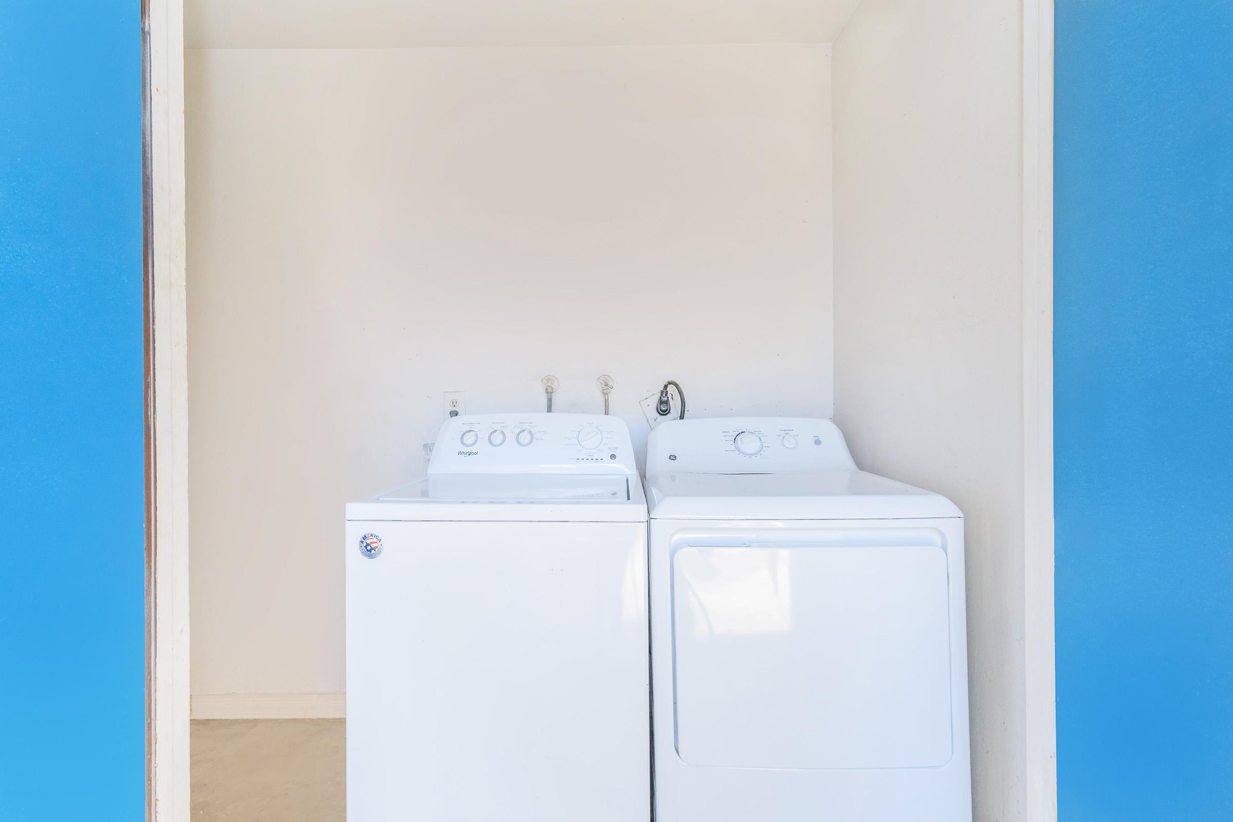 A laundry room featuring a white washing machine and a white dryer side by side against a plain white wall, with light blue walls on either side. The appliances have dials and controls visible, and the floor is light-colored. The space is bright and uncluttered.