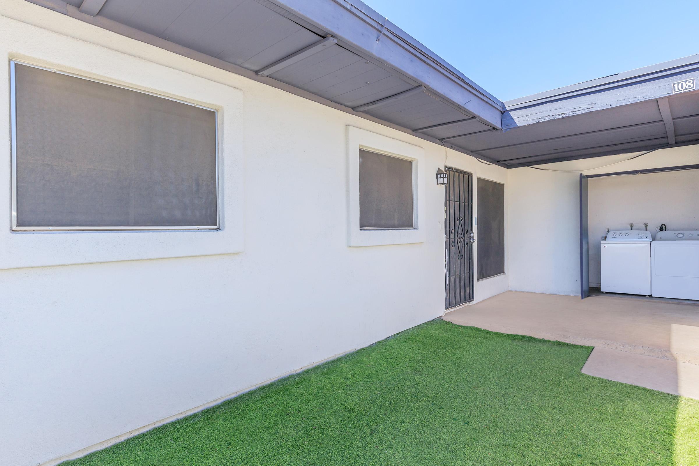 View of a small outdoor area with a green artificial lawn, adjacent to a white wall building. There are two windows with screens, a gate next to the entrance, and a laundry area with a washer and dryer under a shaded roof. The sky is clear, suggesting a sunny day.