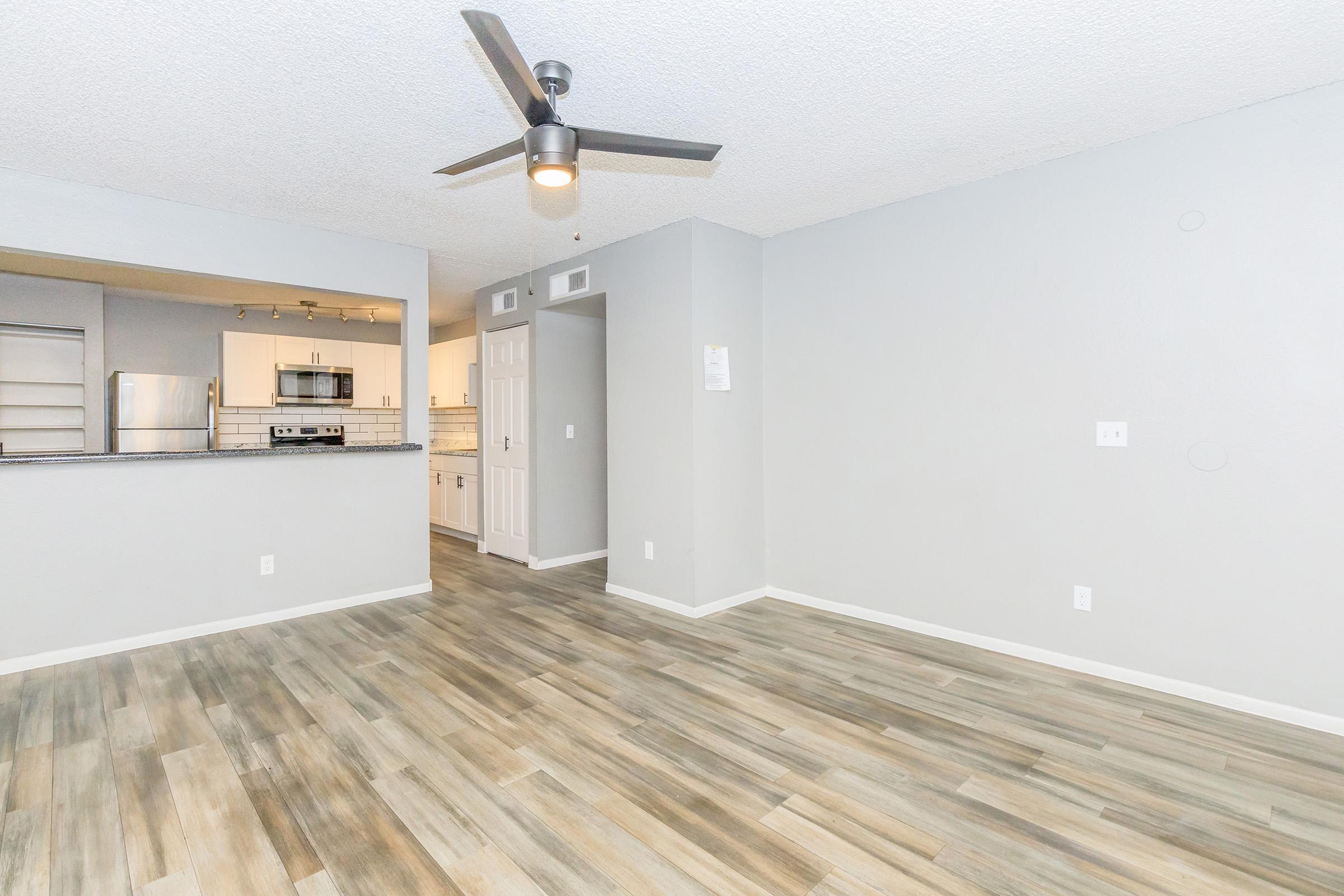 Spacious living area featuring light gray walls, a ceiling fan, and wood-style flooring. A doorway leads to a kitchen with modern appliances in the background. The open layout creates a bright and airy atmosphere, perfect for entertaining or everyday living.