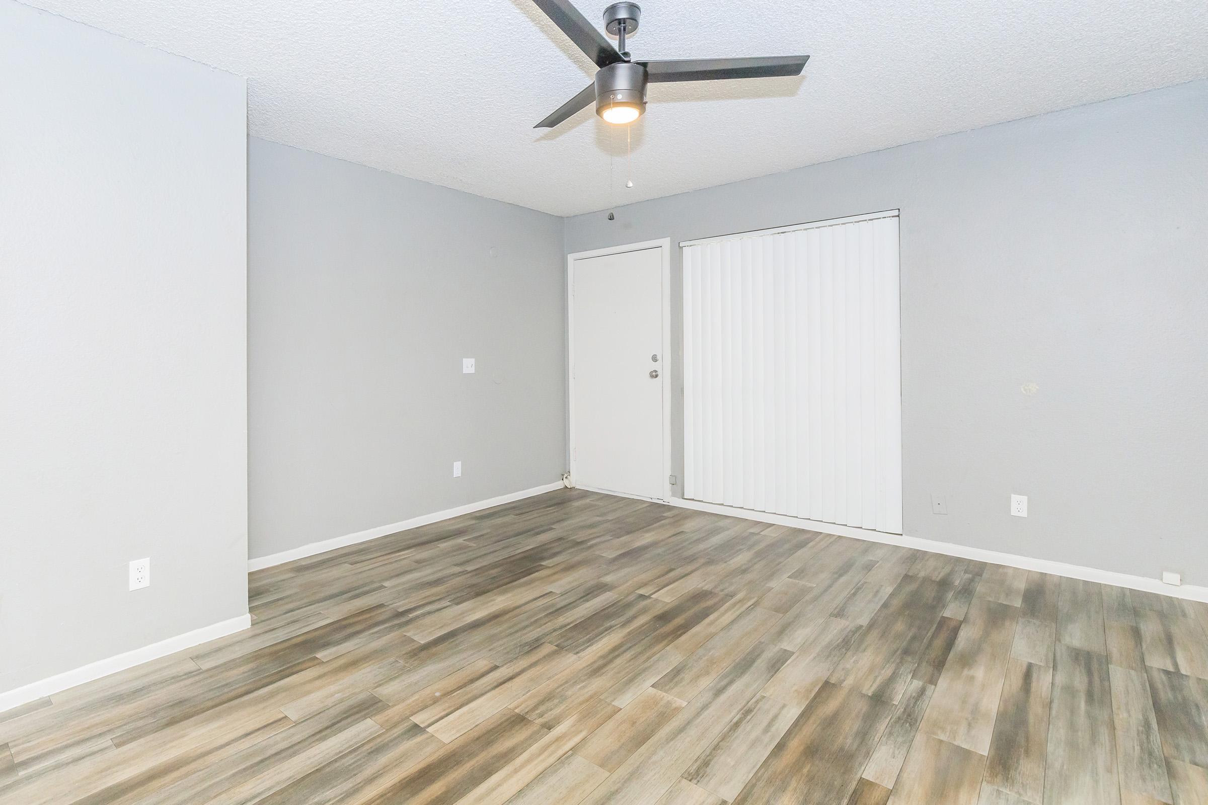 A spacious, minimalistic living room featuring light gray walls, a ceiling fan, and a large window covered with vertical blinds. The flooring is a blend of light and dark wood tiles, creating a modern and open atmosphere. The door to the outside is visible on the left side of the image.