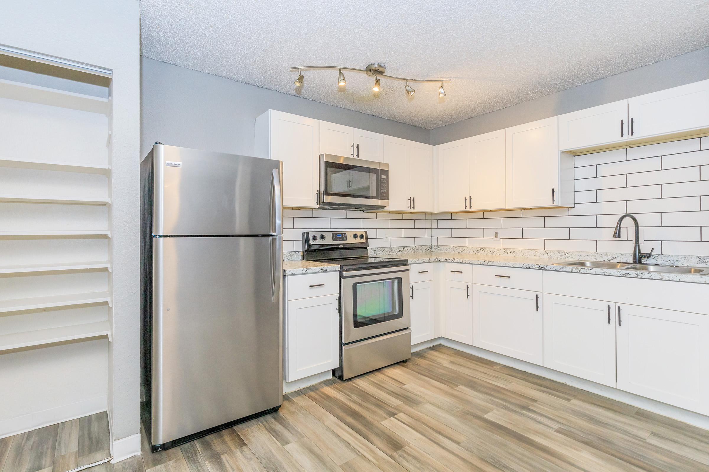 Modern kitchen with stainless steel appliances, including a refrigerator, microwave, and oven. Features white cabinetry, a gray backsplash, and a granite countertop. The floor is finished in light wood tones, and there is ambient lighting from ceiling fixtures. Minimalist design with an open layout.
