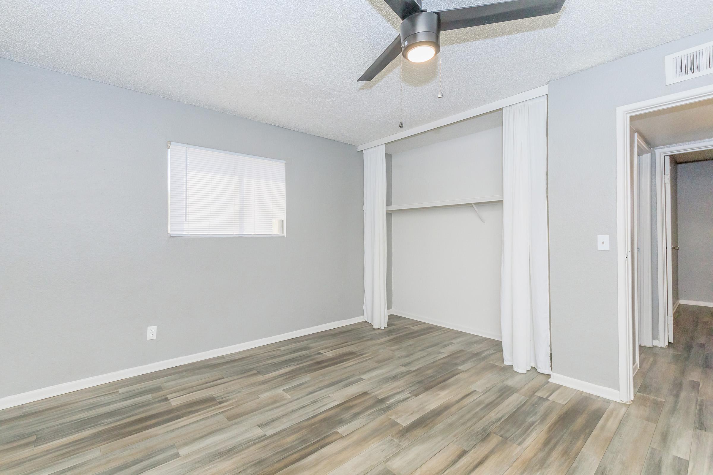 A spacious, well-lit room with light gray walls and wooden vinyl flooring. It features a ceiling fan, a window with blinds, and an open closet with a curtain. The doorway leads to another area, suggesting an efficient layout for an apartment or living space.