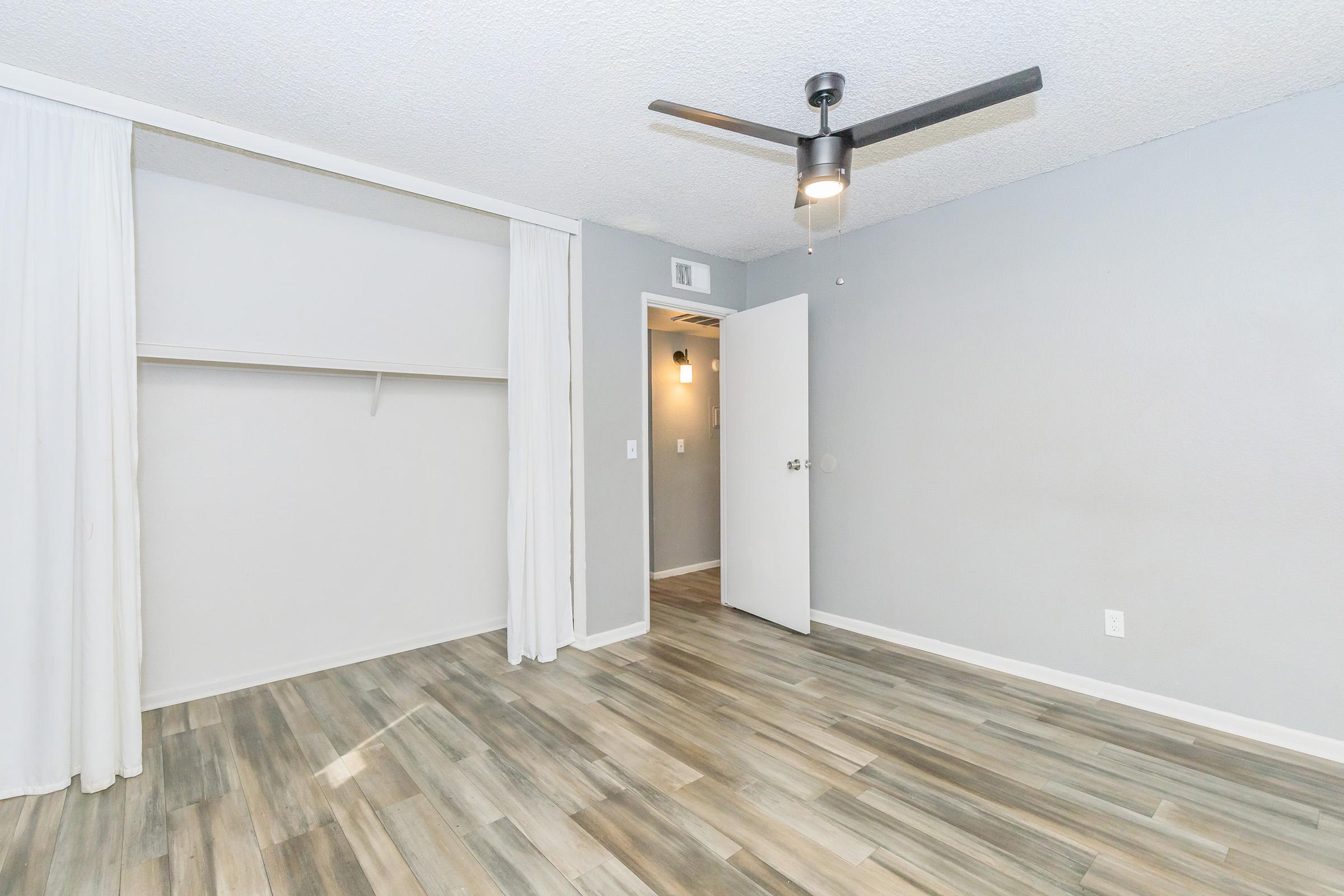 A spacious, unfurnished room featuring light gray walls and a ceiling fan. The floor is made of wood-like tiles. A closet is partially covered by white curtains, and there's a door leading to another space. The overall aesthetic is clean and modern.