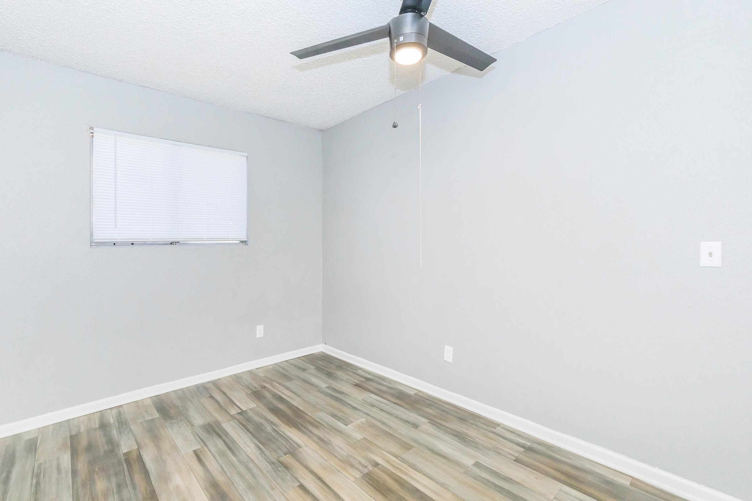 A sparsely furnished room with light gray walls and a wooden floor. There is a small window with blinds on the left side and a ceiling fan above. The space appears clean and minimalist, suitable for various uses.
