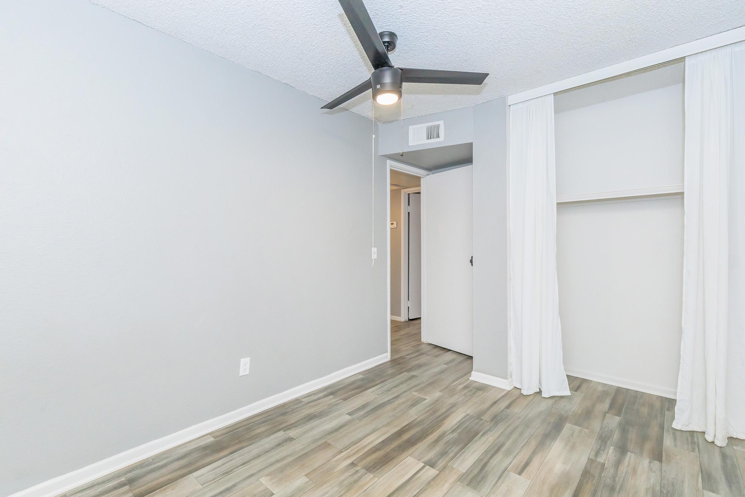 A spacious, empty room with light gray walls and wood-like laminate flooring. A ceiling fan is mounted in the center, and there is a doorway leading to a hallway. On one side, a closet with no doors is visible, adding to the open feel of the space. Natural light enhances the bright ambiance.