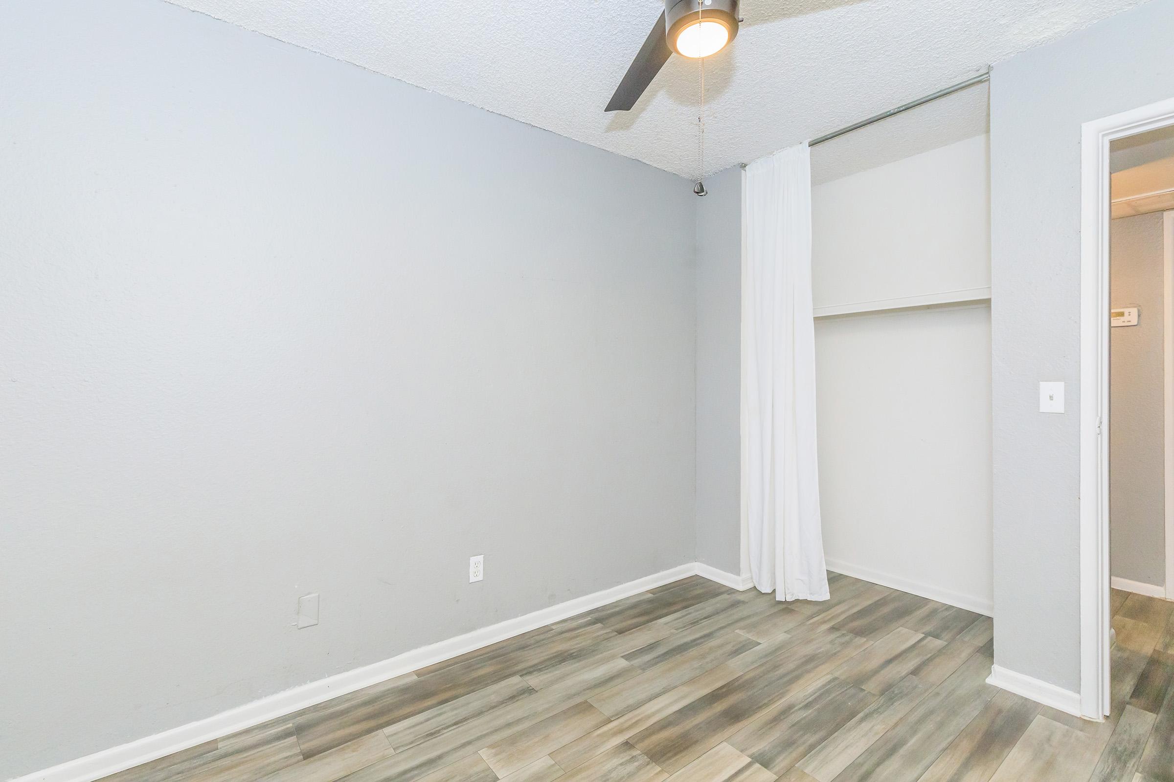 A simple, vacant room featuring light gray walls, a ceiling fan, and wood-like flooring. There is a closet with a white curtain and an interior door on the right side. The overall ambience is neutral and minimalist, making it suitable for various uses.