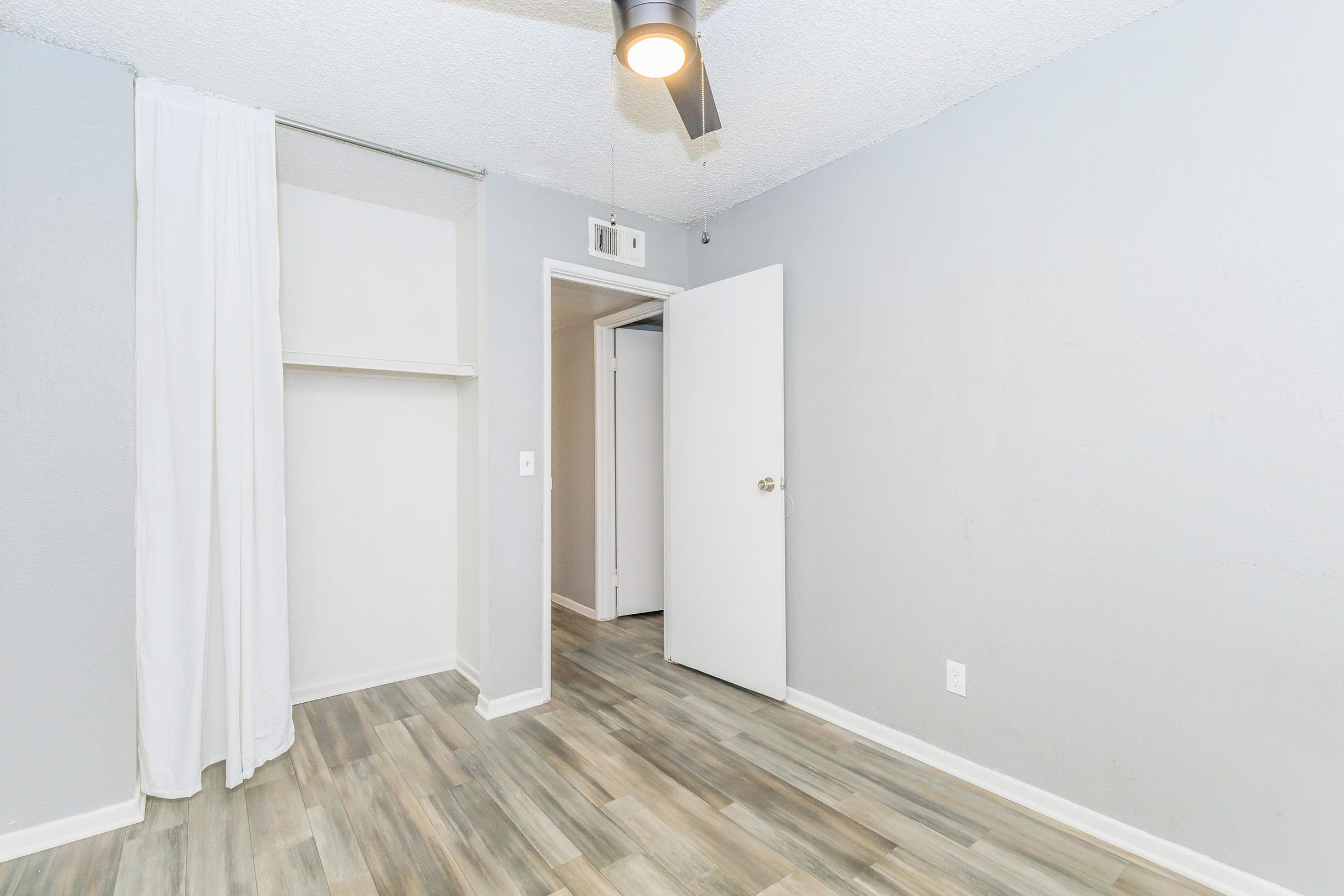 A small, undecorated room with light gray walls and wood-look tile flooring. There is a closet with a white curtain on one side and a doorway leading to another room. A ceiling fan is visible, and the space is well-lit with natural light. The overall atmosphere is clean and minimalistic.