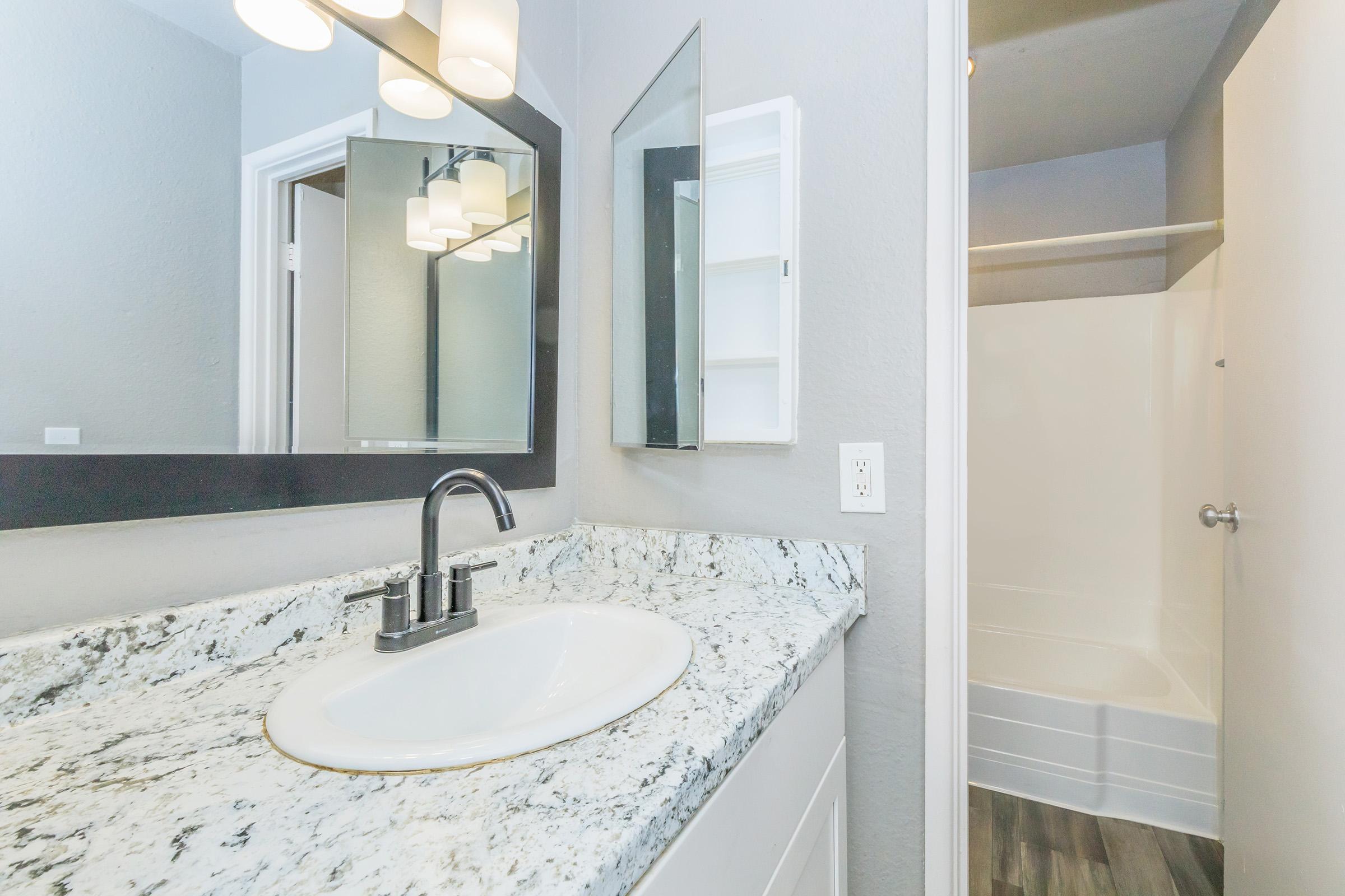 A modern bathroom featuring a granite countertop with a white sink, a large mirror mounted above the sink, a wall-mounted storage cabinet, and a shower area visible through an open door. The walls are painted a soft gray, with stylish light fixtures illuminating the space.