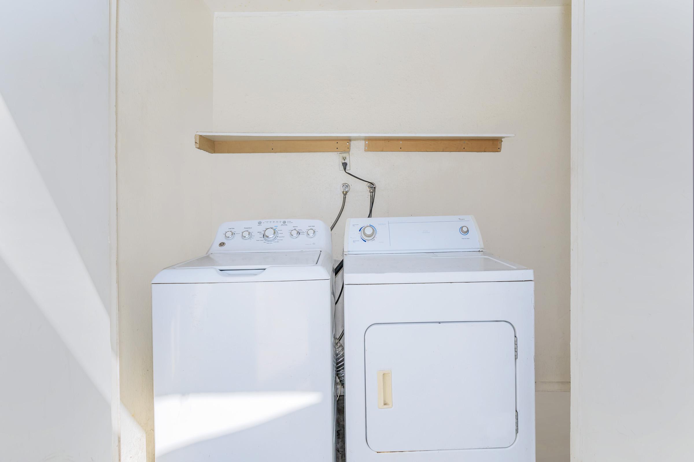 A laundry room featuring a white top-loading washing machine and a white dryer, positioned side by side. Above them is a simple wooden shelf. The walls are painted a light color, creating a clean and spacious appearance. Natural light is visible, highlighting the appliances.
