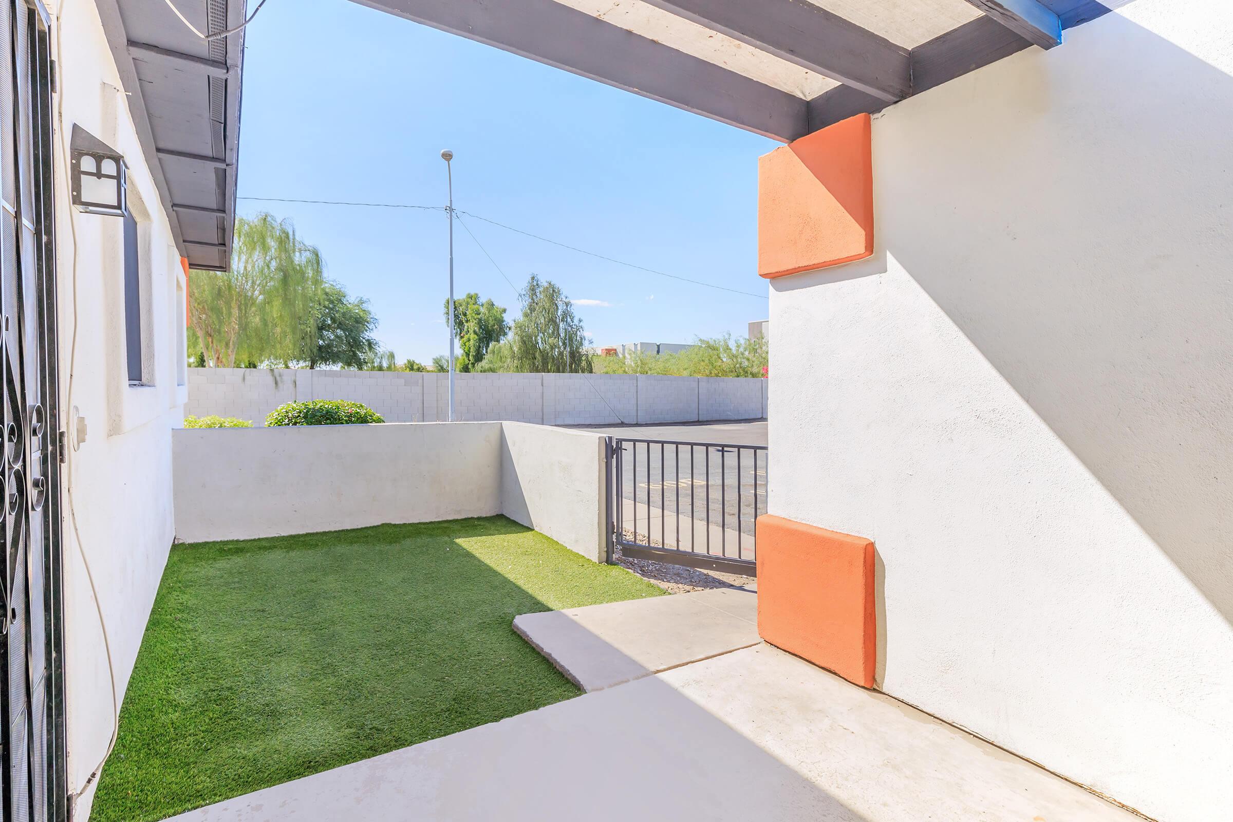 A view of a modern outdoor area featuring artificial grass, a concrete pathway, and a fence. The scene is bright with blue skies and minimal landscaping, highlighting a clean, contemporary design. Two orange wall accents add a pop of color against the white walls.