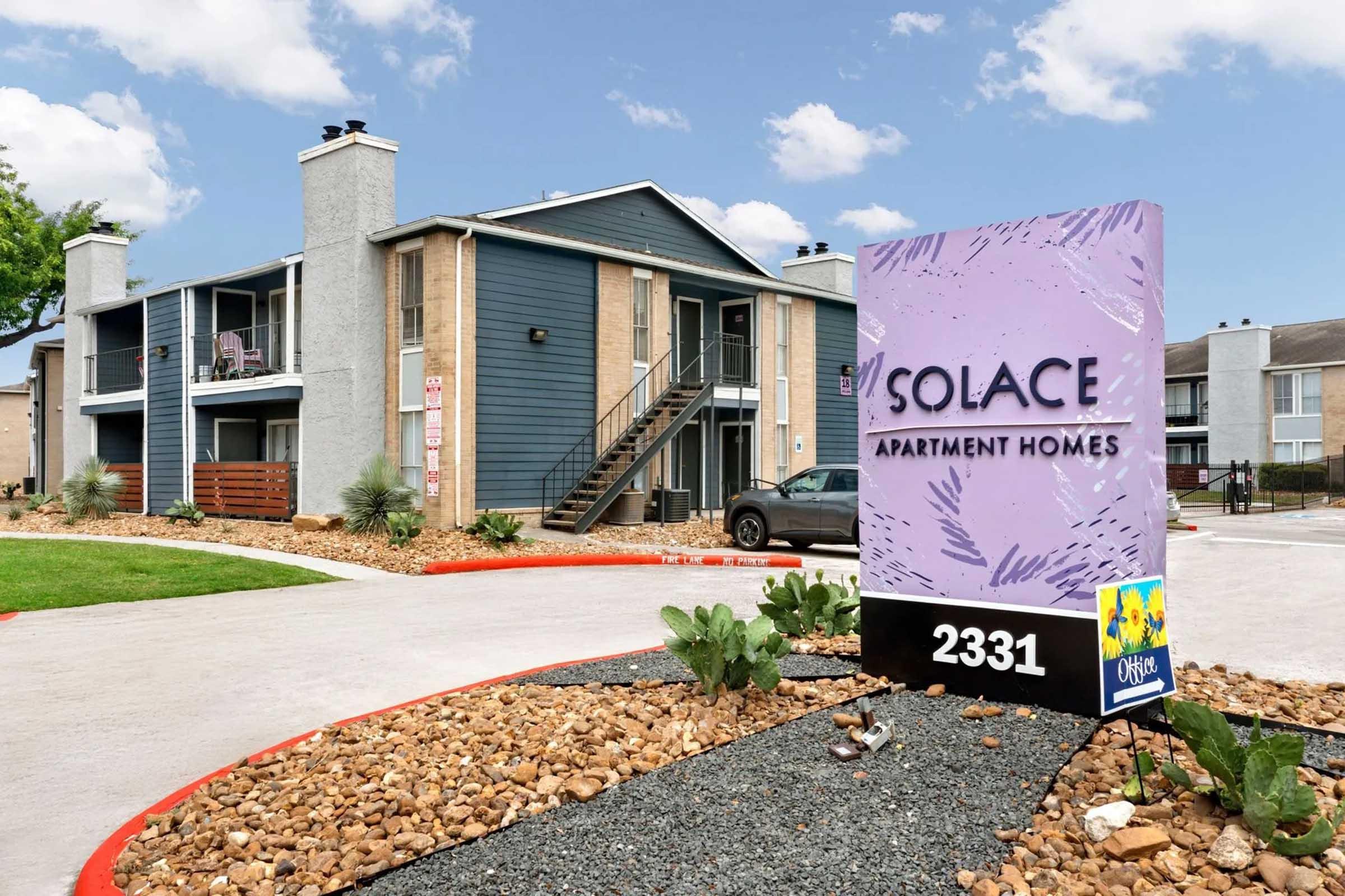 A modern apartment complex named "Solace Apartment Homes" with a welcoming sign in front. The building features a mix of blue and beige exterior, surrounded by well-maintained landscaping, gravel, and potted plants. A parked car is visible in the lot, and the sky is partly cloudy.