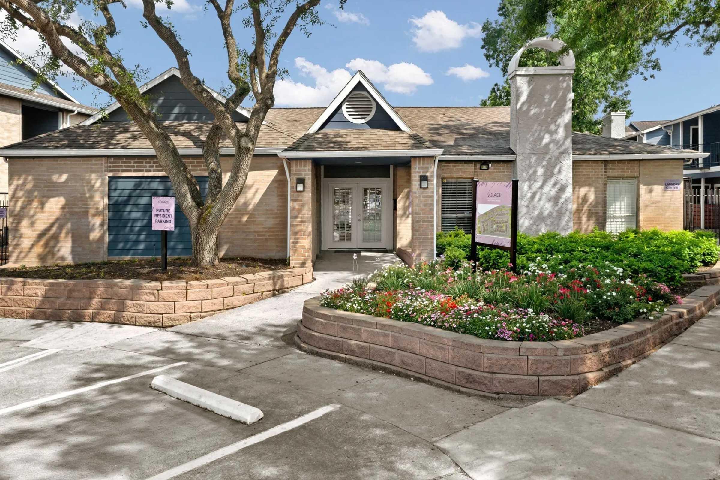 A brick building with a sloped roof, featuring two front doors and framed by trees. Flower beds with colorful blooms surround the entrance, and there are two signs visible. The pathway is paved, leading up to the entrance, which is bright and welcoming. Clear blue sky in the background.