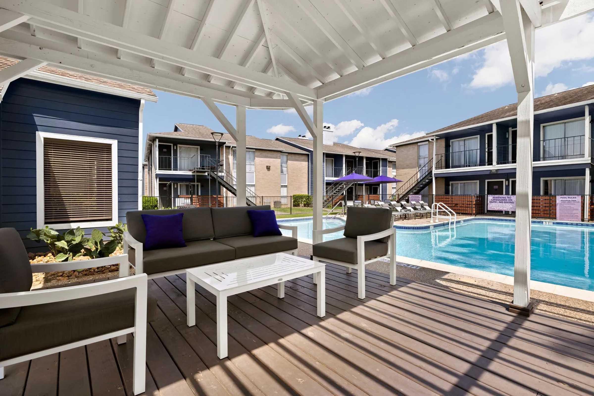 A shaded outdoor seating area with modern furniture beside a swimming pool. The pool is surrounded by multi-story buildings, and there's a purple umbrella providing shade. Sunlight casts shadows on the wooden deck, creating a relaxed atmosphere perfect for lounging.