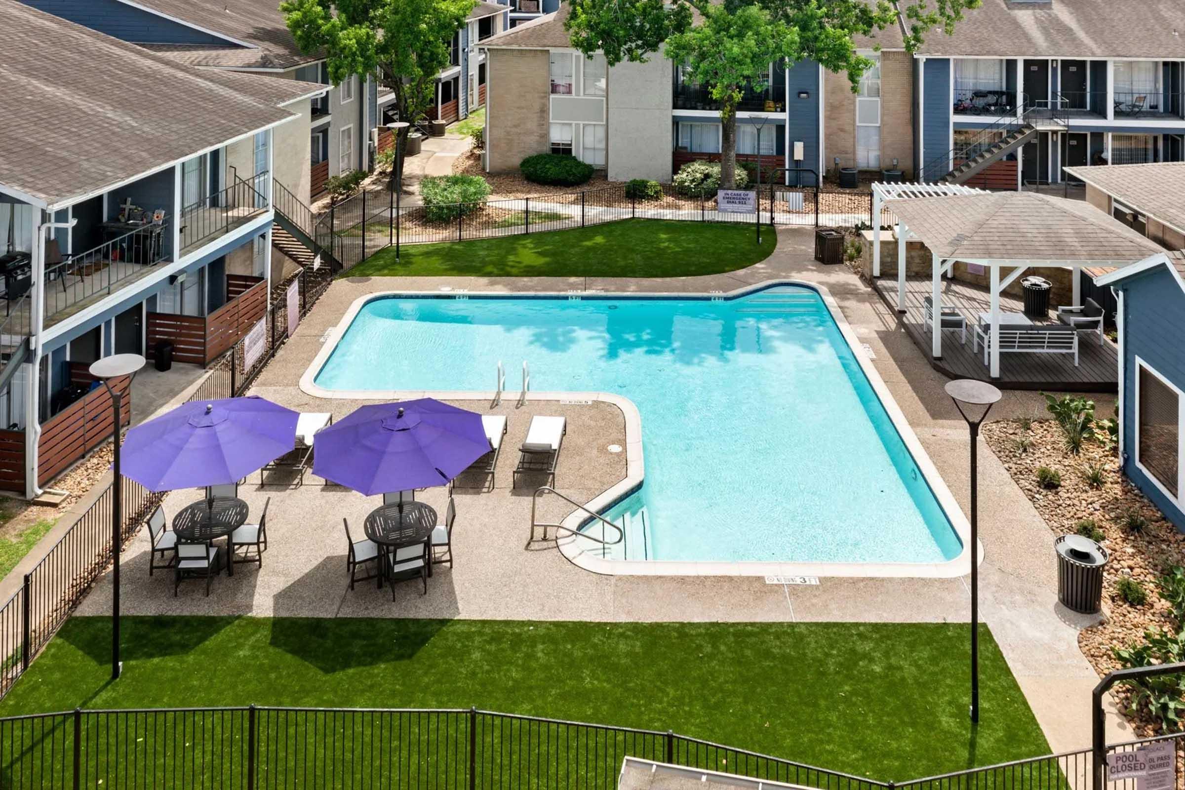 Aerial view of an apartment complex with a swimming pool surrounded by grassy areas. The pool features lounge chairs and two purple umbrellas. Nearby, there are outdoor seating areas and a gazebo. The buildings are two stories tall and have balconies.