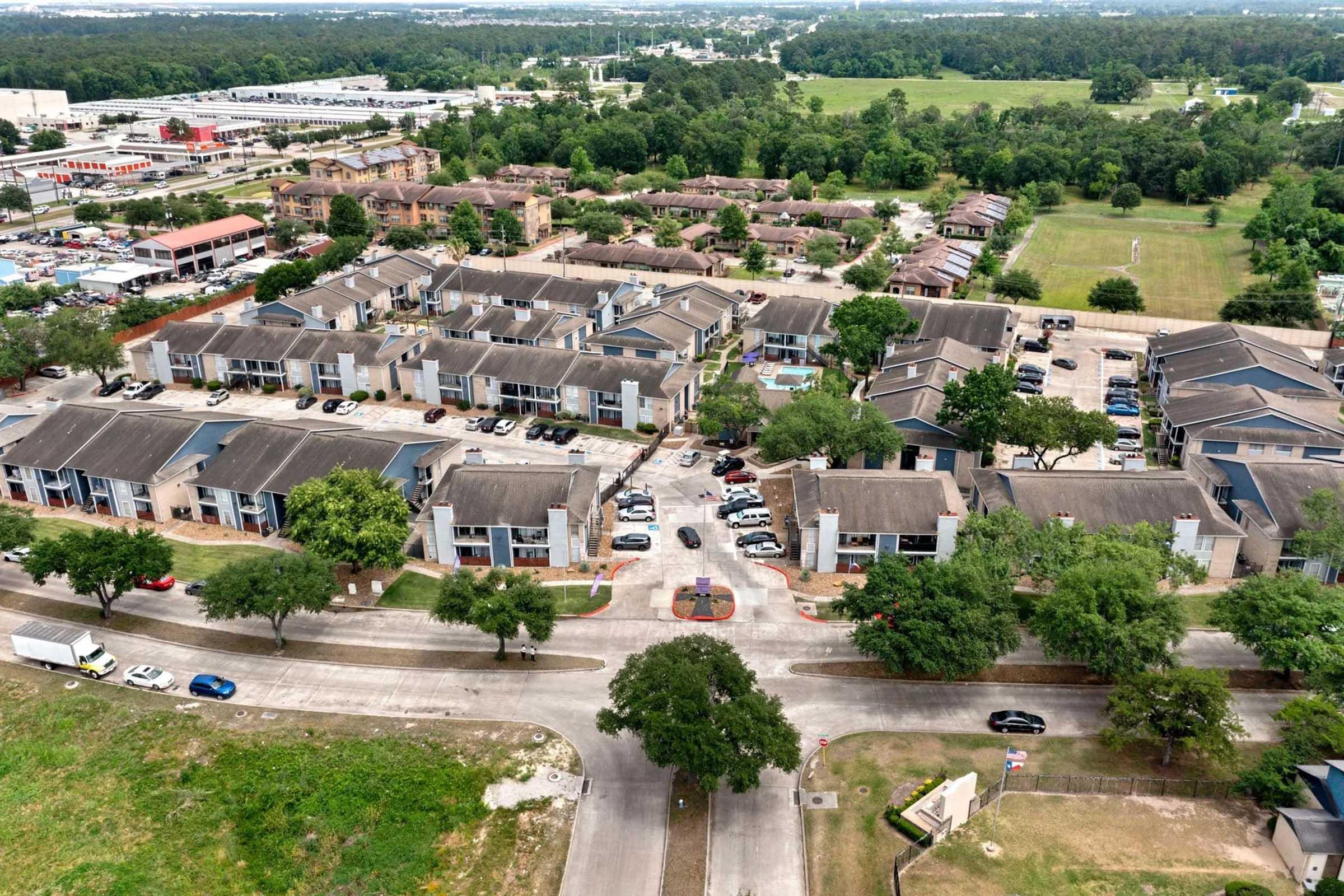 Aerial view of a residential neighborhood featuring multiple low-rise buildings with cars parked along the streets. Green lawns and trees are visible, along with some commercial buildings in the background. The image shows a mix of residential and open green spaces.
