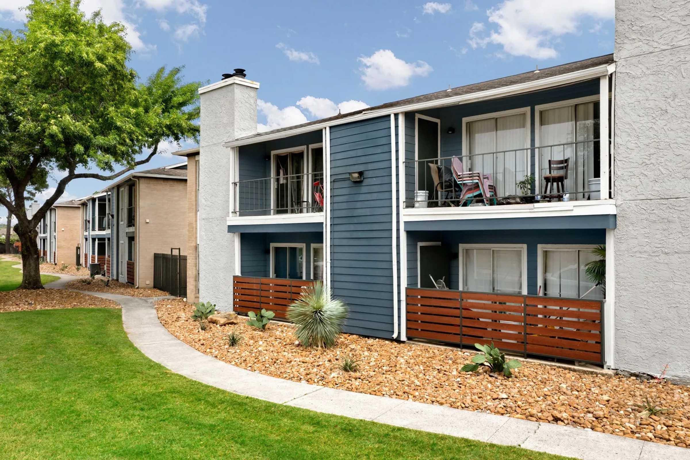 Modern apartment buildings with balconies and greenery. The buildings feature a mix of blue and light gray exteriors, with stone pathways and landscaped areas. Potted plants and lawn accents enhance the outdoor space, creating a welcoming environment under a partly cloudy sky.