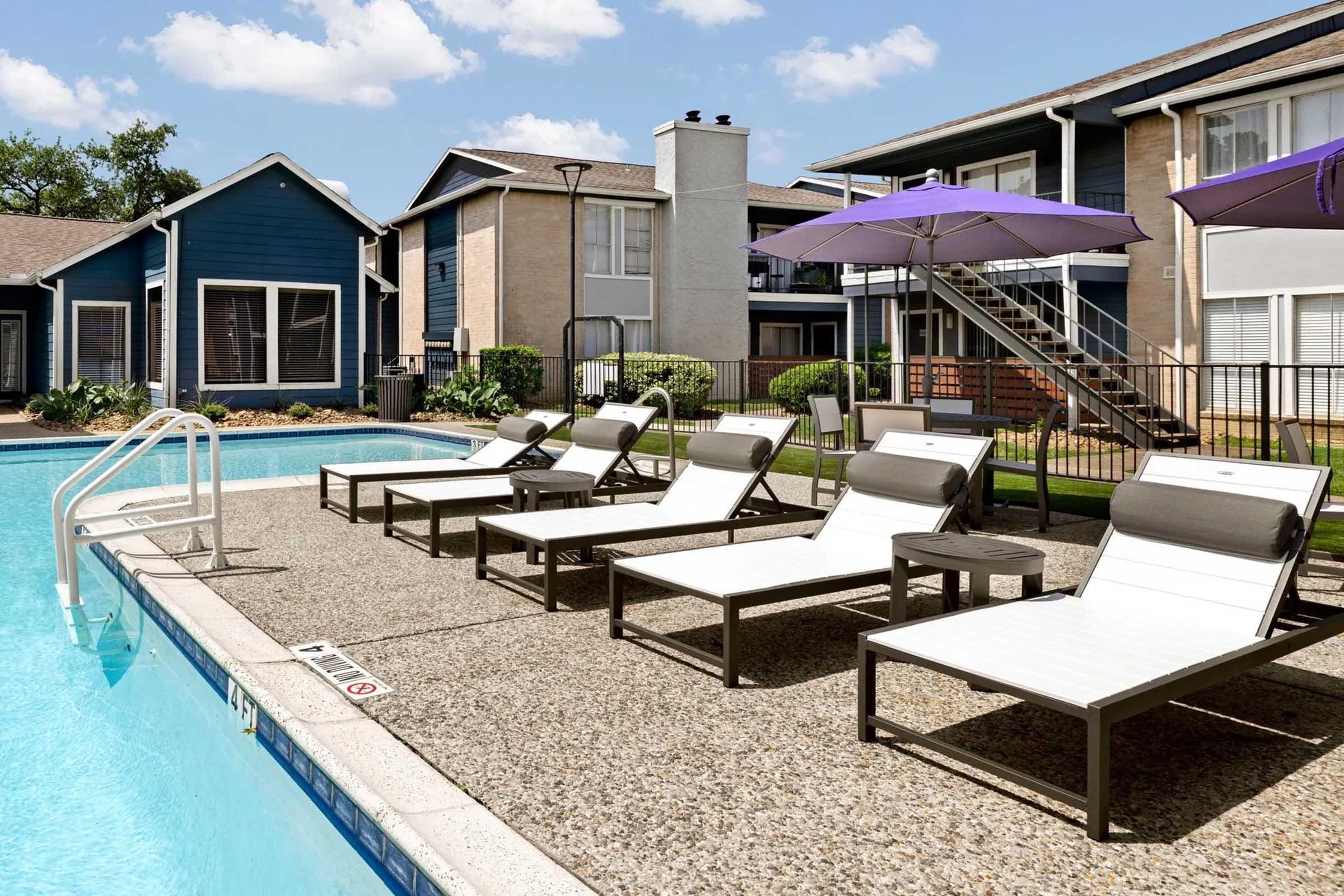 A sunny pool area featuring several sleek lounge chairs beside a clear blue swimming pool, with purple umbrellas providing shade. Surrounding the pool are landscaped gardens and modern apartment buildings in the background, creating a relaxing outdoor atmosphere.