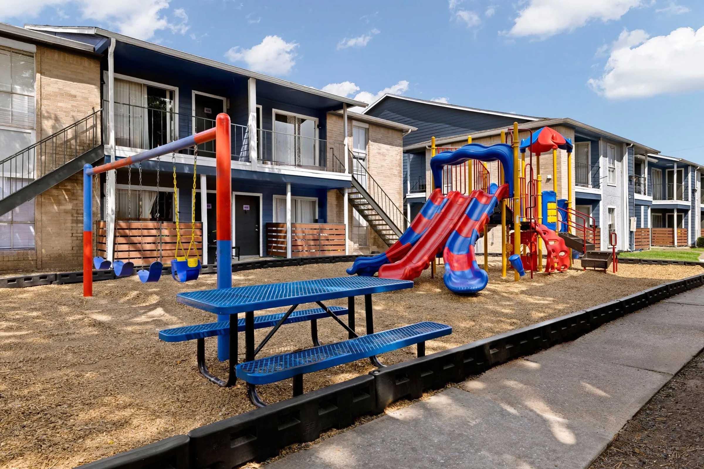 A colorful playground featuring a slide, climbing structure, and swings, located in a residential area with apartment buildings in the background. The ground is covered with gravel, and there is a blue picnic table nearby, providing a space for children to play and socialize.