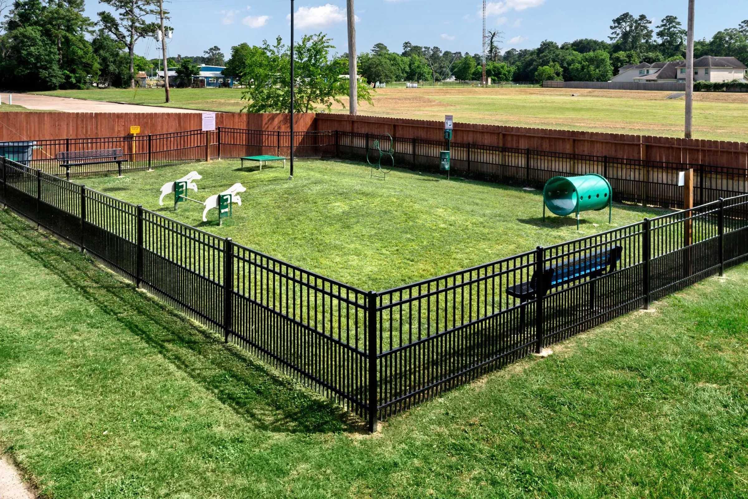 A fenced dog park featuring green grass, playground equipment for dogs, and benches for owners. There are several agility structures, such as tunnels and jumps, along with plenty of open space for dogs to play. The park is surrounded by a wooden fence and offers a sunny outdoor environment.