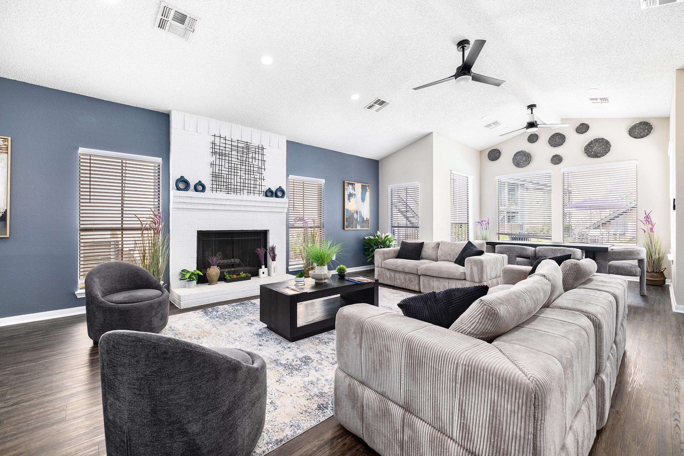 A stylish living room featuring a modern design with a neutral color palette. It includes a light gray sectional sofa, a dark coffee table, and plush armchairs. The walls are accented in dark blue, and decorative elements like plants and wall art add a contemporary touch. Large windows provide natural light.