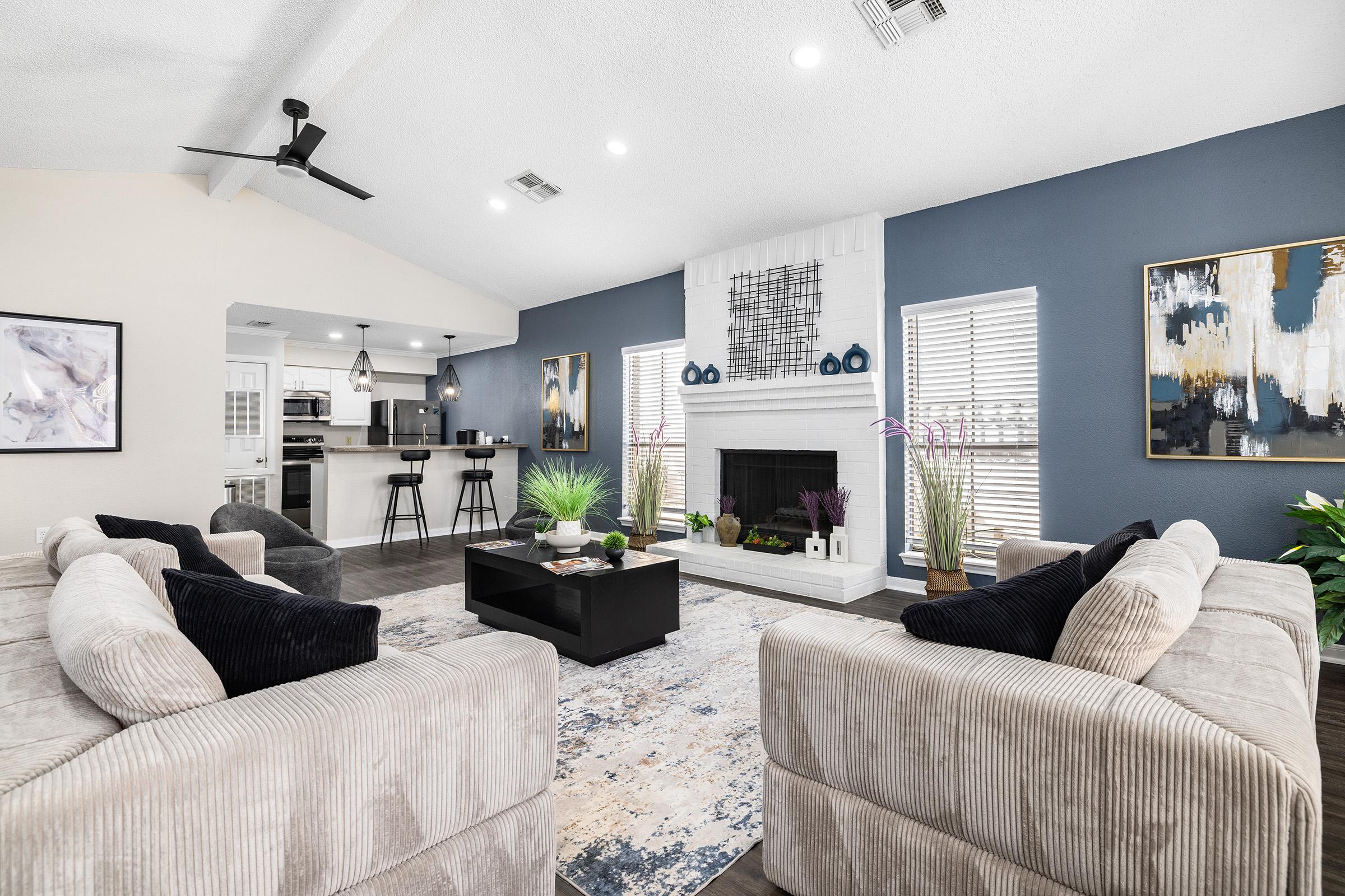 A modern, open-concept living room featuring a white fireplace and large windows, adorned with plants and decorative artwork. Two plush sofas face a central coffee table on a colorful area rug, with a dining area visible in the background, characterized by stylish bar stools and contemporary decor.