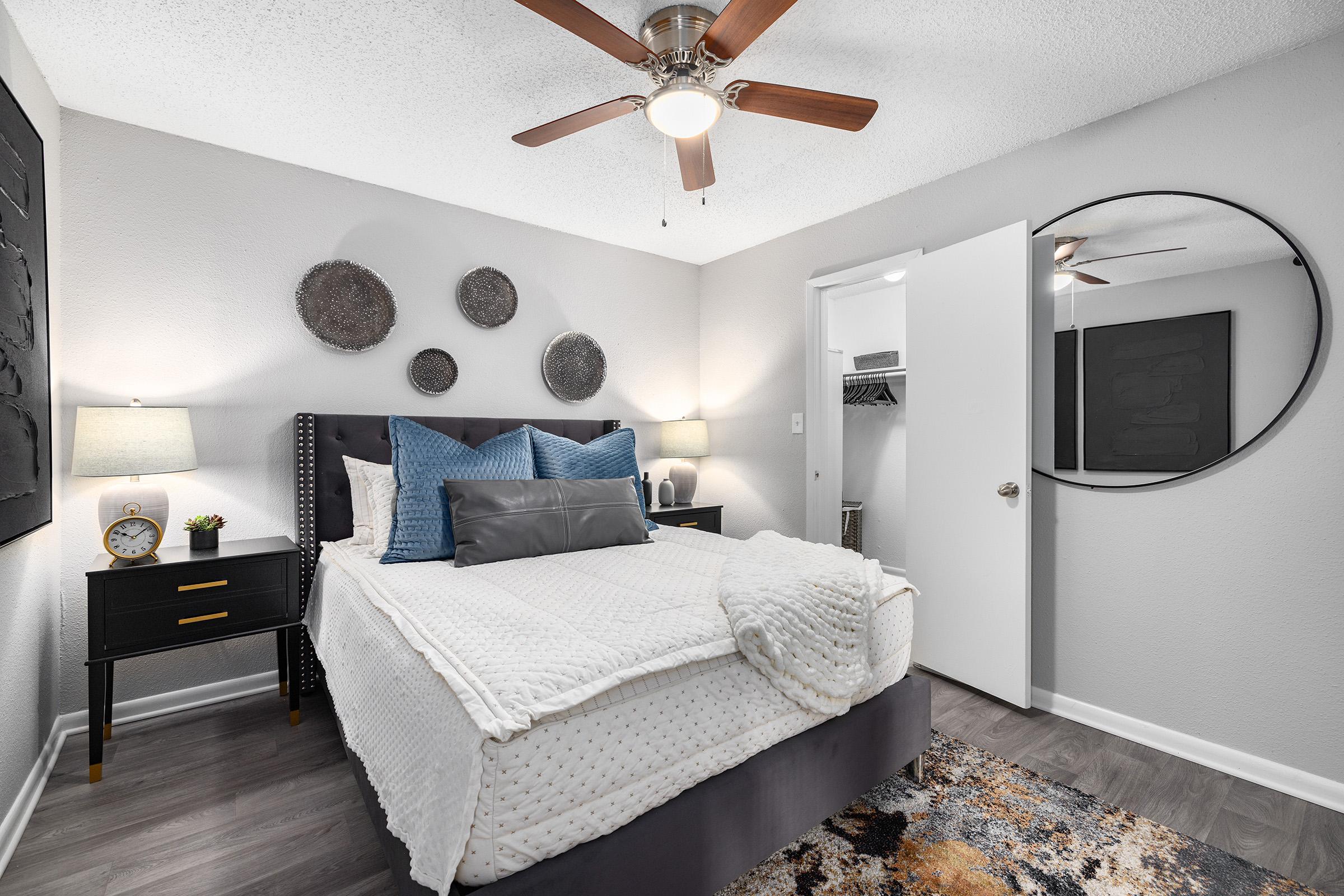 A cozy bedroom featuring a queen-sized bed with a white quilt, fluffy pillows, and a decorative gray throw. Beside the bed are two modern nightstands with lamps. A round mirror hangs on the wall, and there's a closet door open in the background. The room has a stylish rug and wall art, creating a welcoming ambiance.