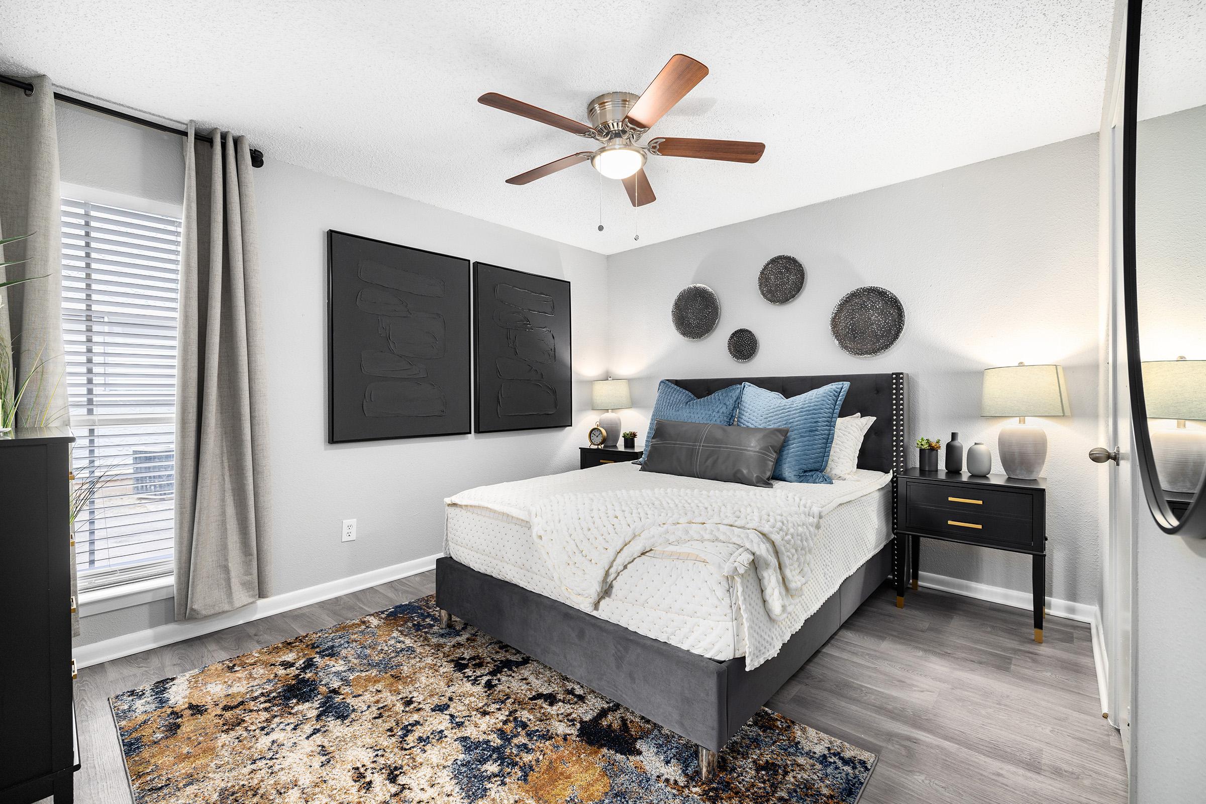 A modern bedroom featuring a neatly made bed with white and blue bedding. There are two bedside lamps on black nightstands, decorative wall art, and a ceiling fan. A large mirror reflects the room, while a colorful area rug adds warmth to the space. Bright natural light enters through the window, illuminating the decor.
