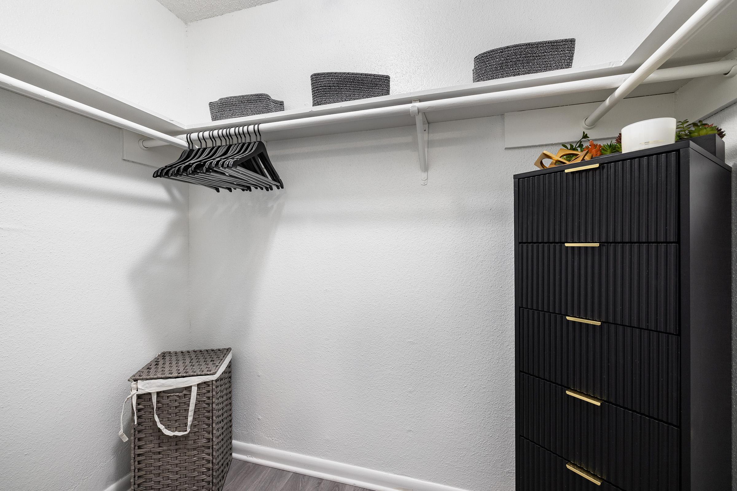 A well-organized closet featuring a row of hangers, storage baskets on a shelf, a tall black cabinet with multiple drawers, and a wicker laundry basket. The walls are painted white, and the floor is a light wood finish.