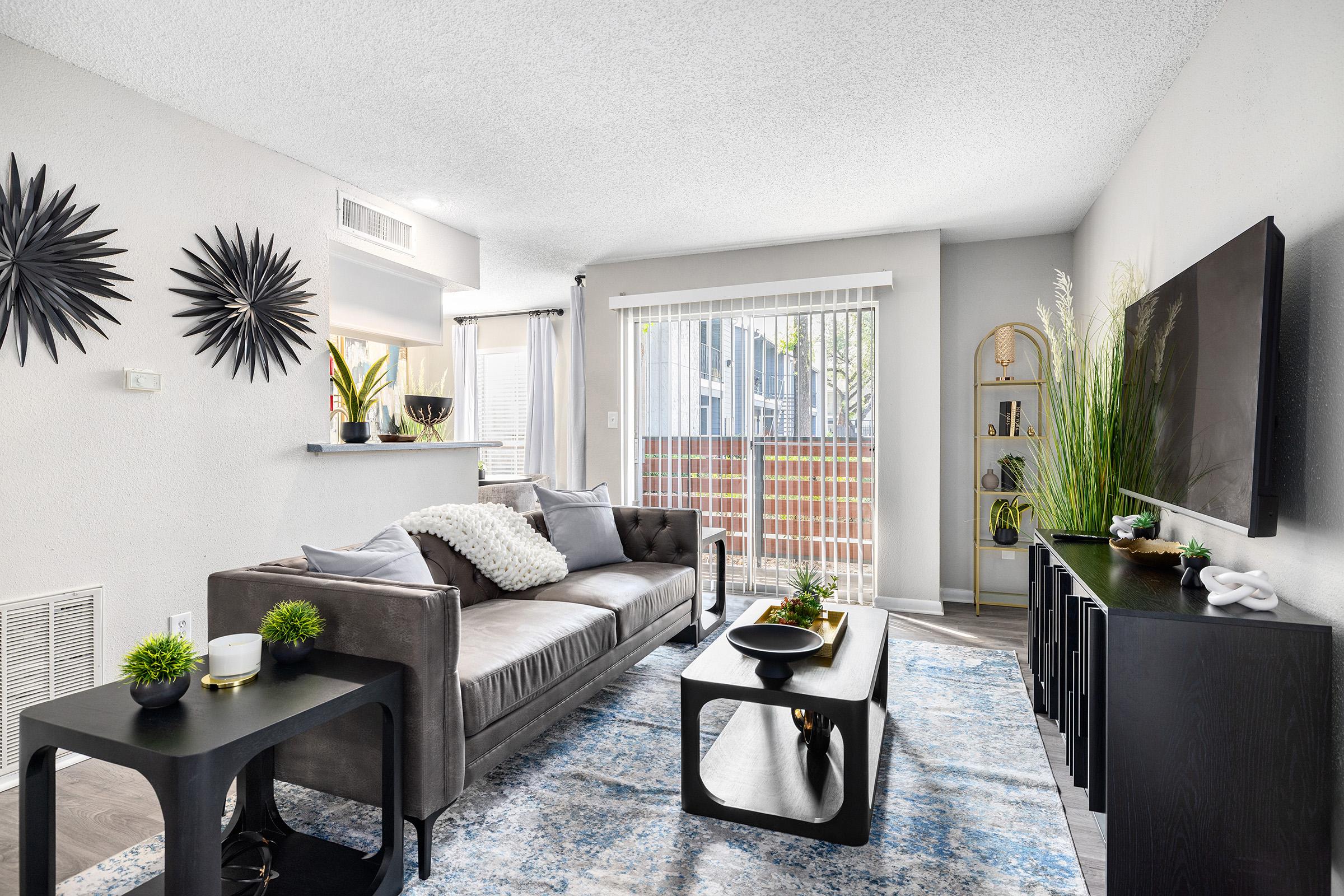 A modern living room featuring a gray sofa adorned with decorative pillows, a black coffee table, and a stylish TV stand. The space is complemented by a vibrant area rug, potted plants, and wall decor, including star-shaped mirrors. Natural light streams through sliding glass doors, enhancing the cozy atmosphere.