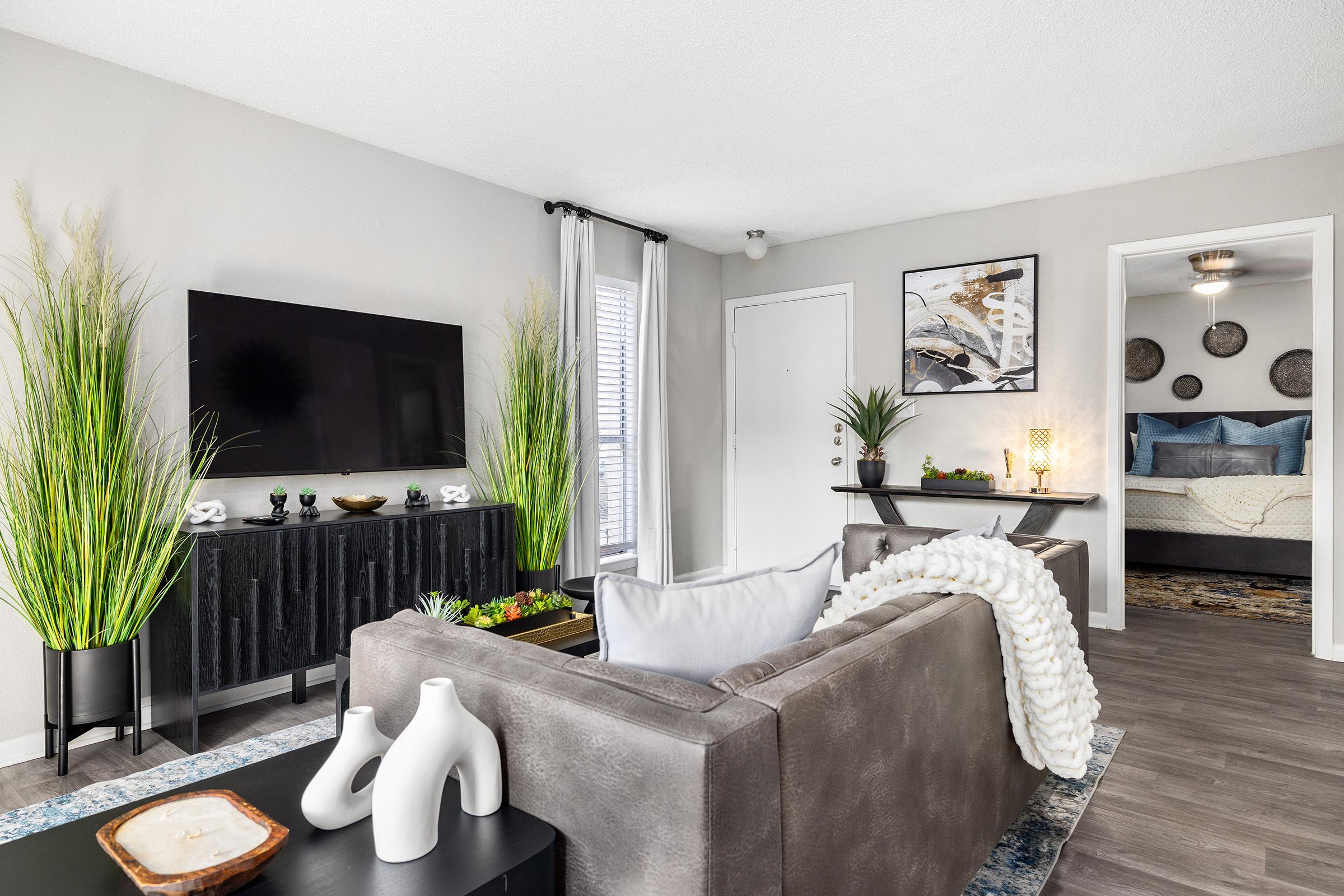 A modern living room featuring a comfortable gray sofa with decorative pillows, a black media console with a flat-screen TV, and two large green plants. The space showcases light grey walls, a colorful area rug, and a doorway leading to a bedroom with a neutral color scheme and art on the walls.