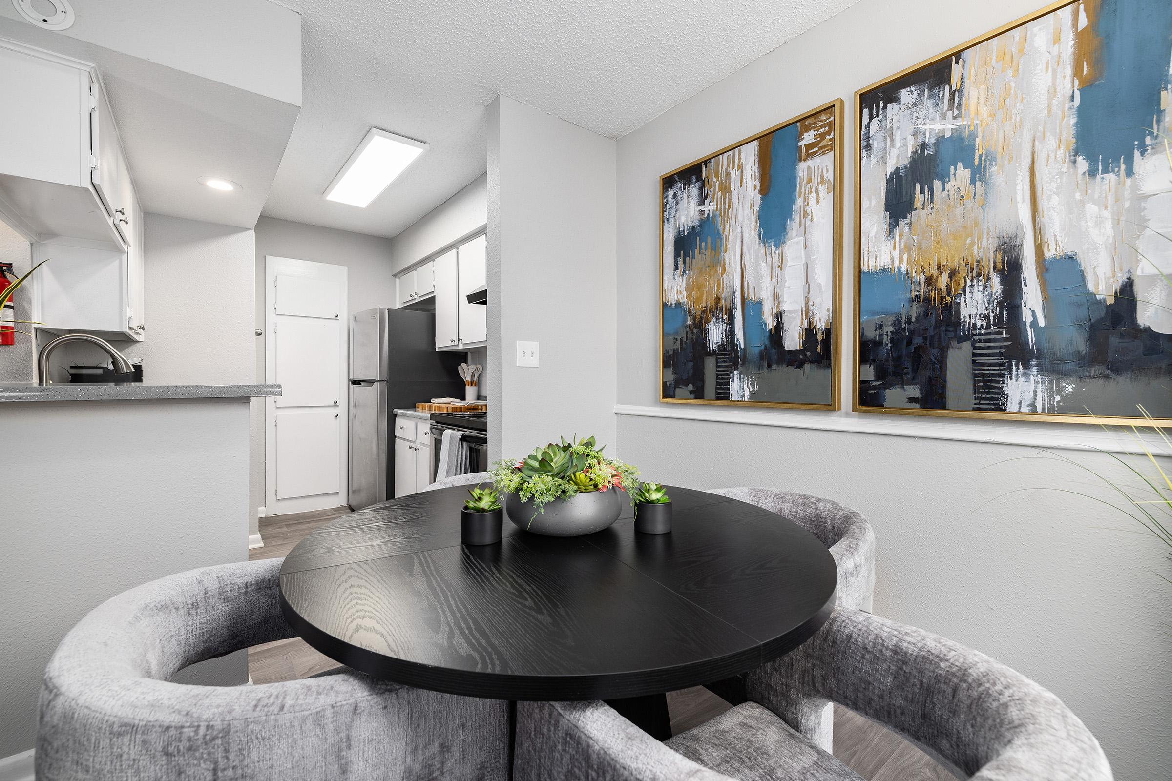 A modern dining area featuring a round black table with plush gray chairs. Two abstract paintings adorn the wall, showcasing blue and gold tones. In the background, a sleek kitchen is visible, with white cabinetry and stainless steel appliances. A small plant arrangement centers the table.