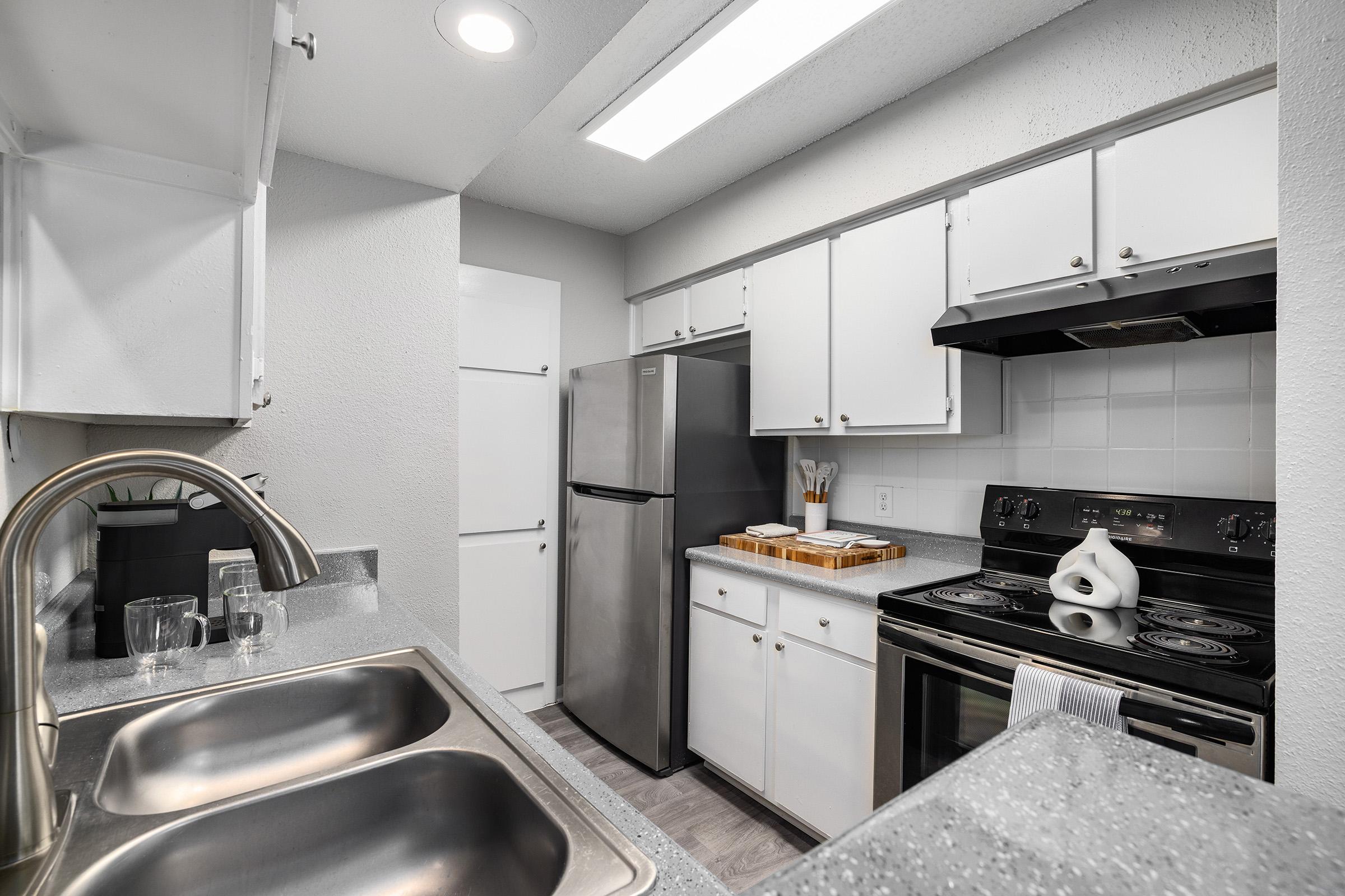 A modern kitchen featuring white cabinets, stainless steel refrigerator, and oven. A double sink with a brushed metal faucet is visible, along with a wooden cutting board on the counter. The walls are light-colored, and there's an overhead light fixture illuminating the space.