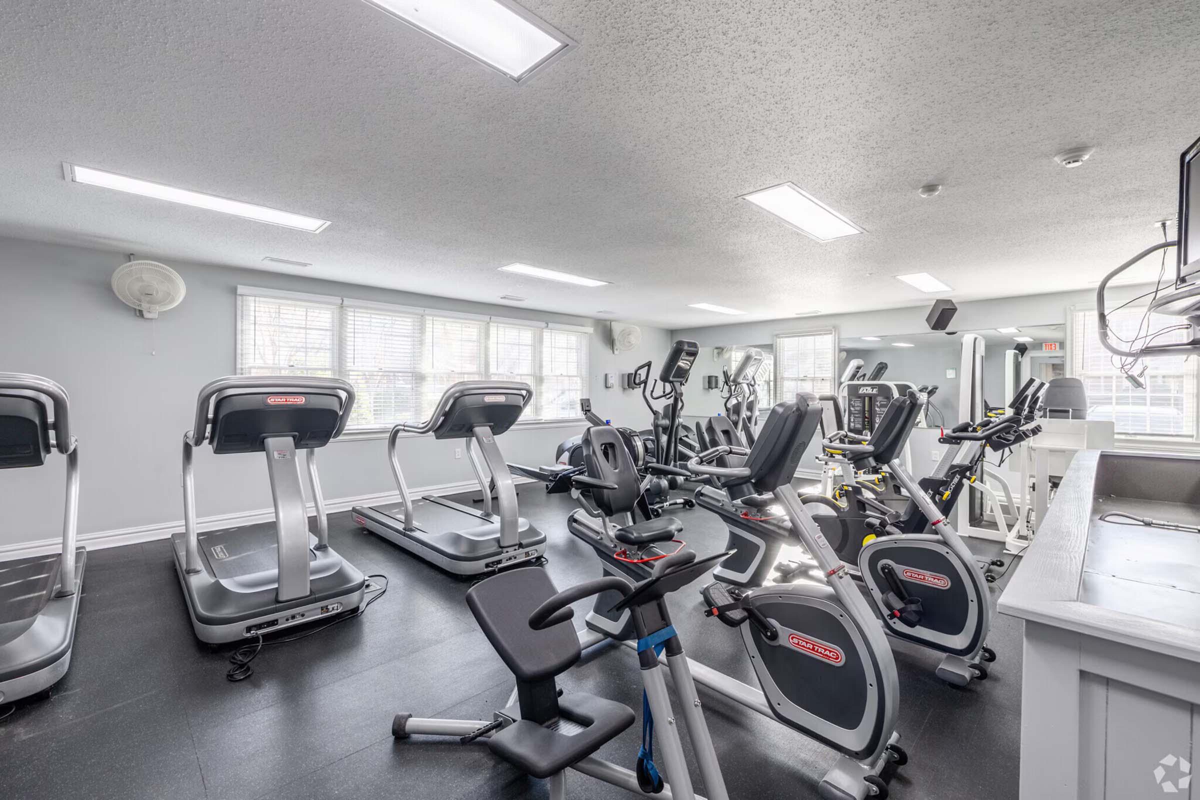A modern gym interior featuring various exercise equipment, including treadmills, stationary bikes, and elliptical machines. Large windows provide natural light, and the space has a clean, organized layout with a neutral color palette and bright overhead lighting.